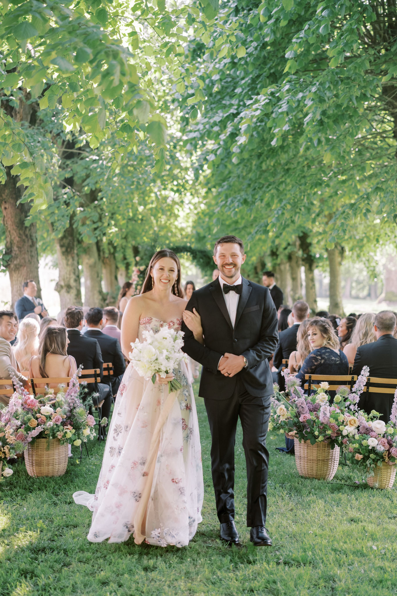 The newlywed couple walks back down the aisle following an outdoor ceremony held beneath a canopied tree-lined alley, with seated guests on wooden benches visible in the background. The bride wears a strapless ivory ball gown with an all-over floral embroidered pattern in blush pink and muted purple tones, and carries a loose bouquet of white sweet peas, ivory ranunculus, and soft white blooms. The groom wears a classic black tuxedo with a black bow tie. Wicker baskets filled with garden-style arrangements of dusty pink roses, blush ranunculus, mauve blooms, and soft peach flowers line both sides of the aisle. The portrait-style shot is taken at mid-distance, capturing the couple from head to toe with approximately 40–50 guests seated behind them in a naturally lit, classic French outdoor setting.