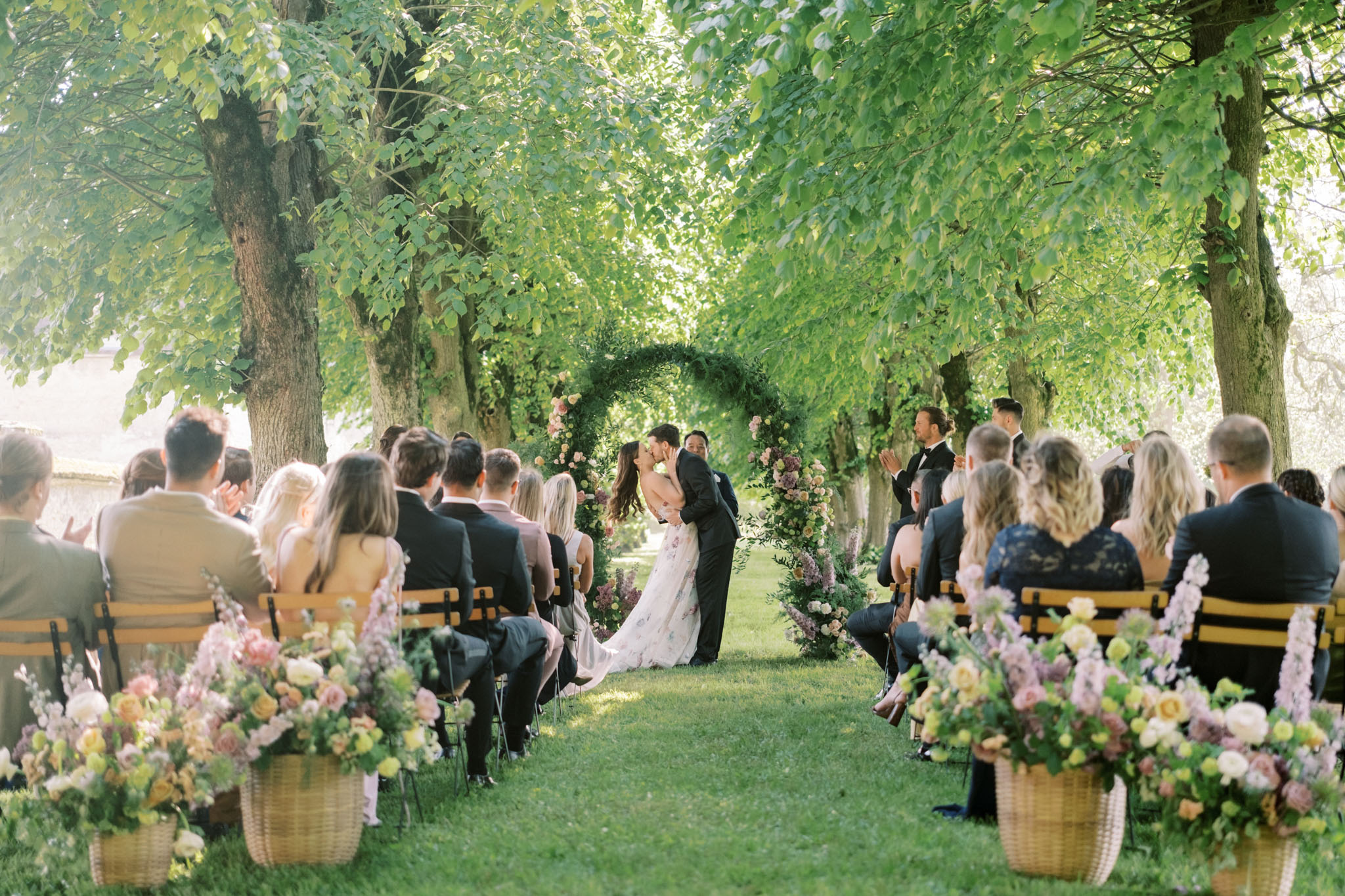Garden ceremony under circular floral arch with blush and mauve blooms wicker aisle baskets and forty guests on wooden ben...