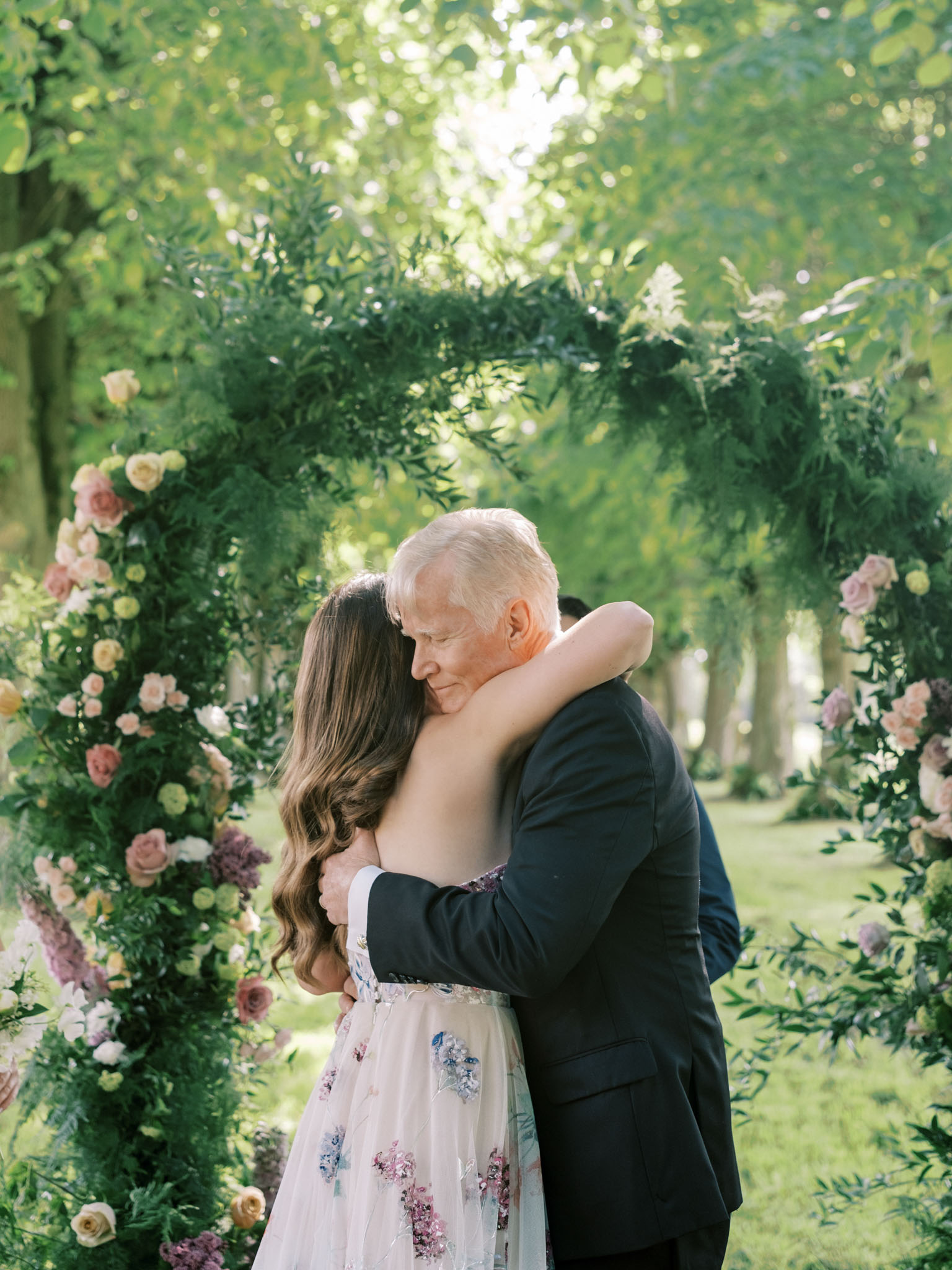 Bride embracing her father at the altar in front of a circular greenery arch adorned with pink mauve and yellow roses