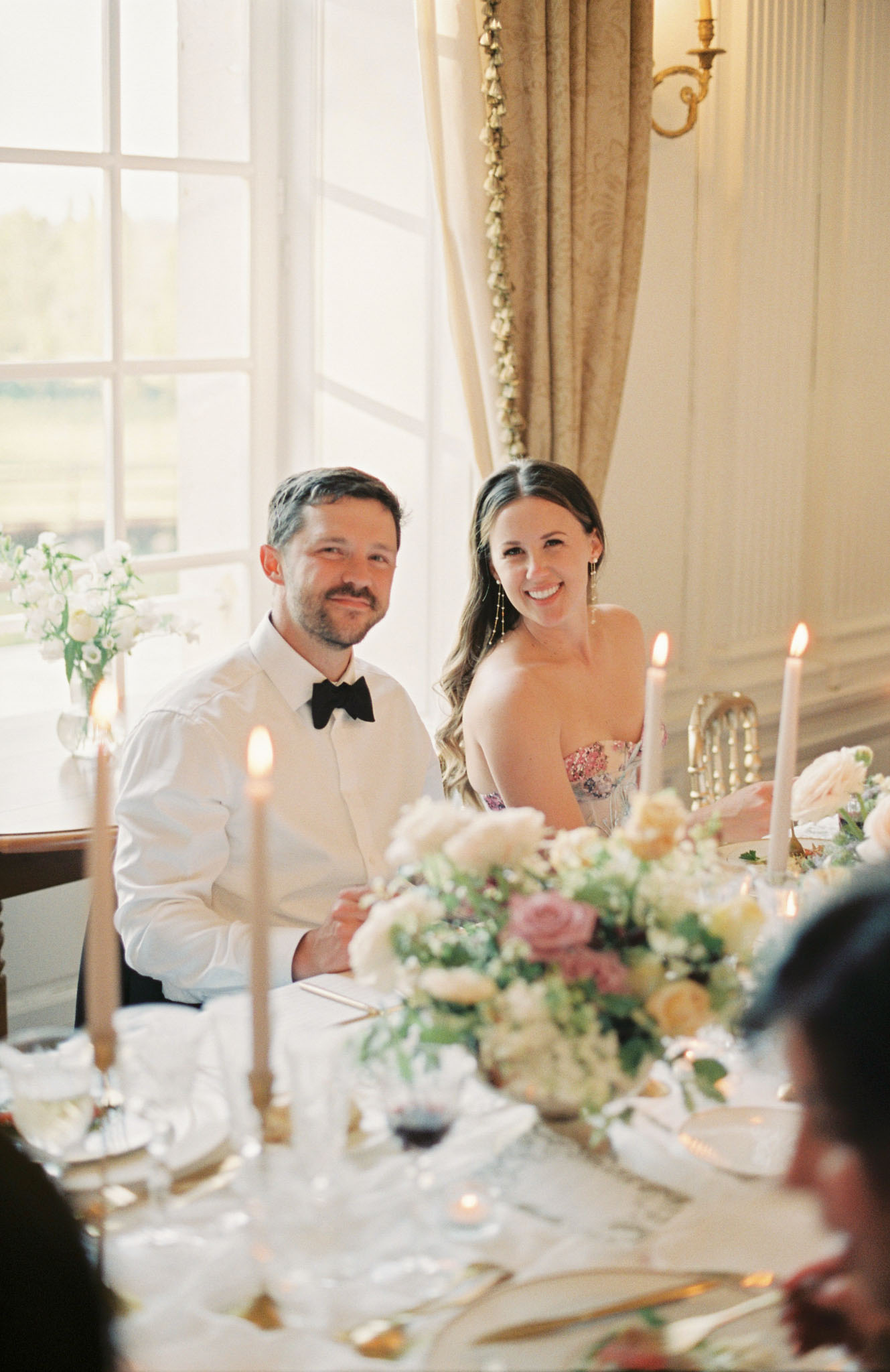 Couple smiling at reception table with gold flatware, crystal glasses, and pink rose centerpiece in chateau dining room