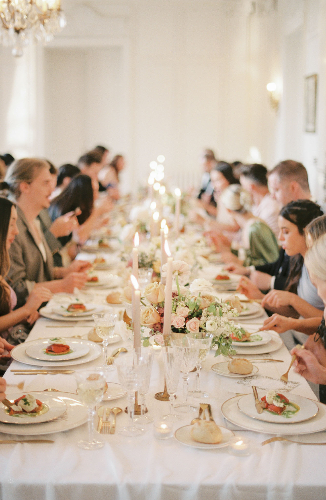 Guests seated at a long banquet table with blush rose centerpieces and taper candles in a white ballroom