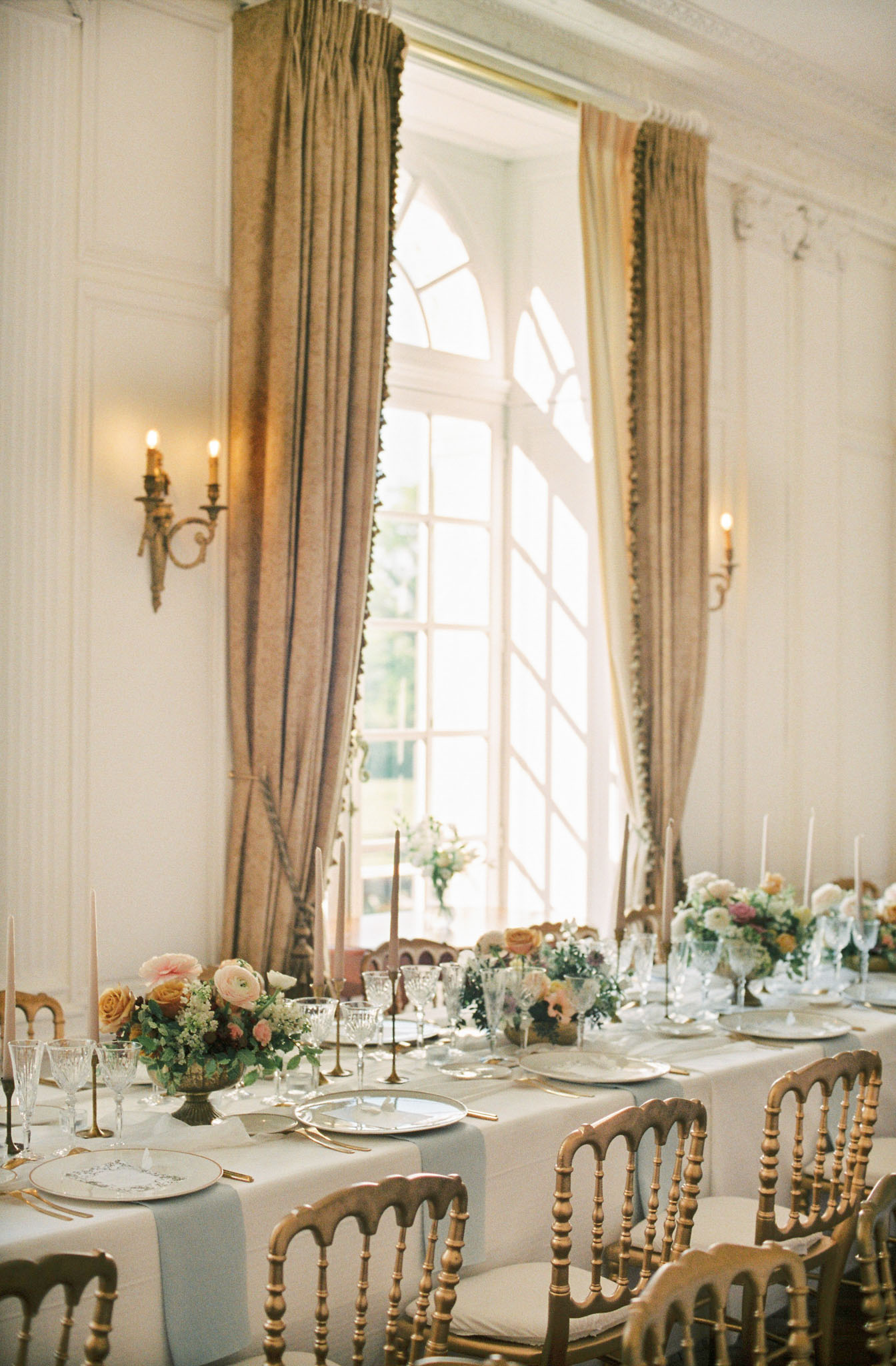 A reception tablescape detail shot taken inside a French château ballroom or salon, with no guests present. The long dining table is dressed in a pale grey-blue linen runner over a white tablecloth, set with gold flatware, ornate white charger plates with calligraphy menus, and crystal glassware. Floral centerpieces in low bronze compote vessels feature blush ranunculus, peach and amber roses, white blooms, and trailing greenery. Slim taper candles in brass candleholders — including both blush pink and dark brown tapers — are spaced along the table. Gold Napoleon-style chairs with ivory seat cushions surround the table. The room features white boiserie wall panelling, gilt bronze double-arm wall sconces with lit candles, and tall arched French windows dressed with floor-length antique gold silk drape curtains with a fringe trim detail. The overall styling palette is soft blush, peach, amber, and muted sage with gold accents, consistent with a classic French romantic aesthetic. Wide shot with a slightly elevated angle. Potential venue feature image.