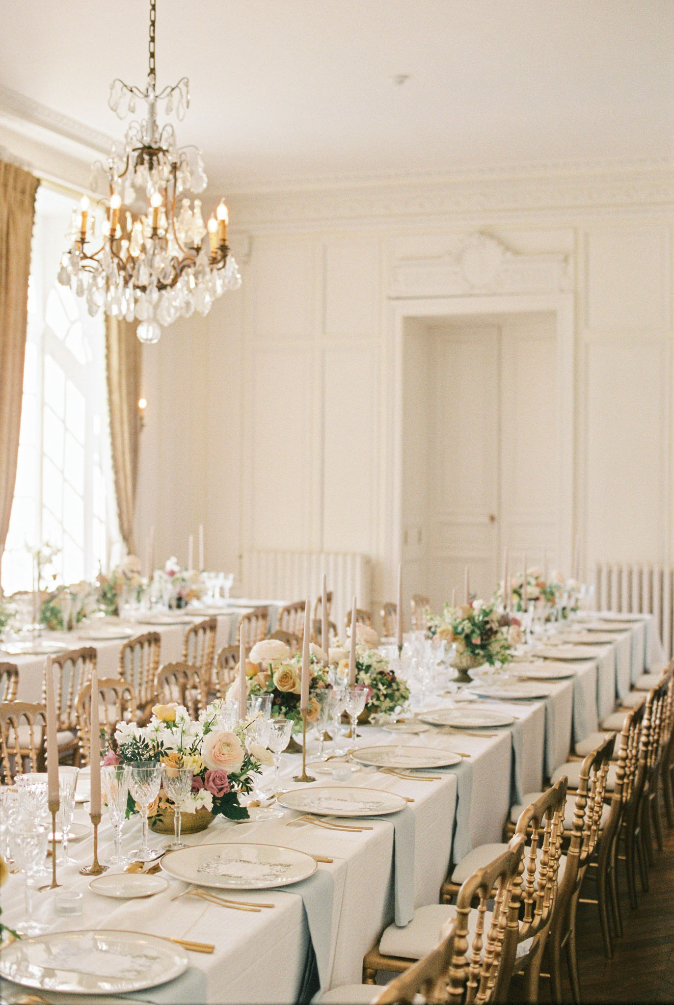 Indoor feasting table with blue-grey linen, gold Chiavari chairs, and pastel florals in chateau