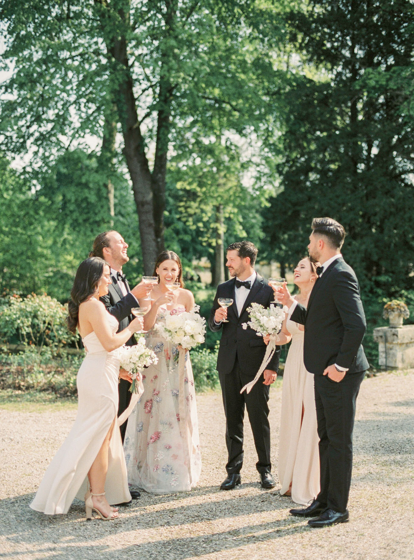 A bridal party of six is gathered outdoors on a gravel path, raising coupe glasses in a champagne toast during what appears to be a cocktail hour. The bride wears a strapless ball gown with an ivory base and an all-over floral embroidery pattern in pink, blue, and green tones, holding a large bouquet of white peonies and garden roses. The two bridesmaids wear blush champagne satin slip-style gowns with thin straps and carry smaller bouquets of white flowers with trailing ivory ribbons. The three groomsmen are dressed in classic black tuxedos with bow ties. The group is laughing and interacting naturally in a medium-distance portrait shot taken on a sunny day in a formal garden setting.