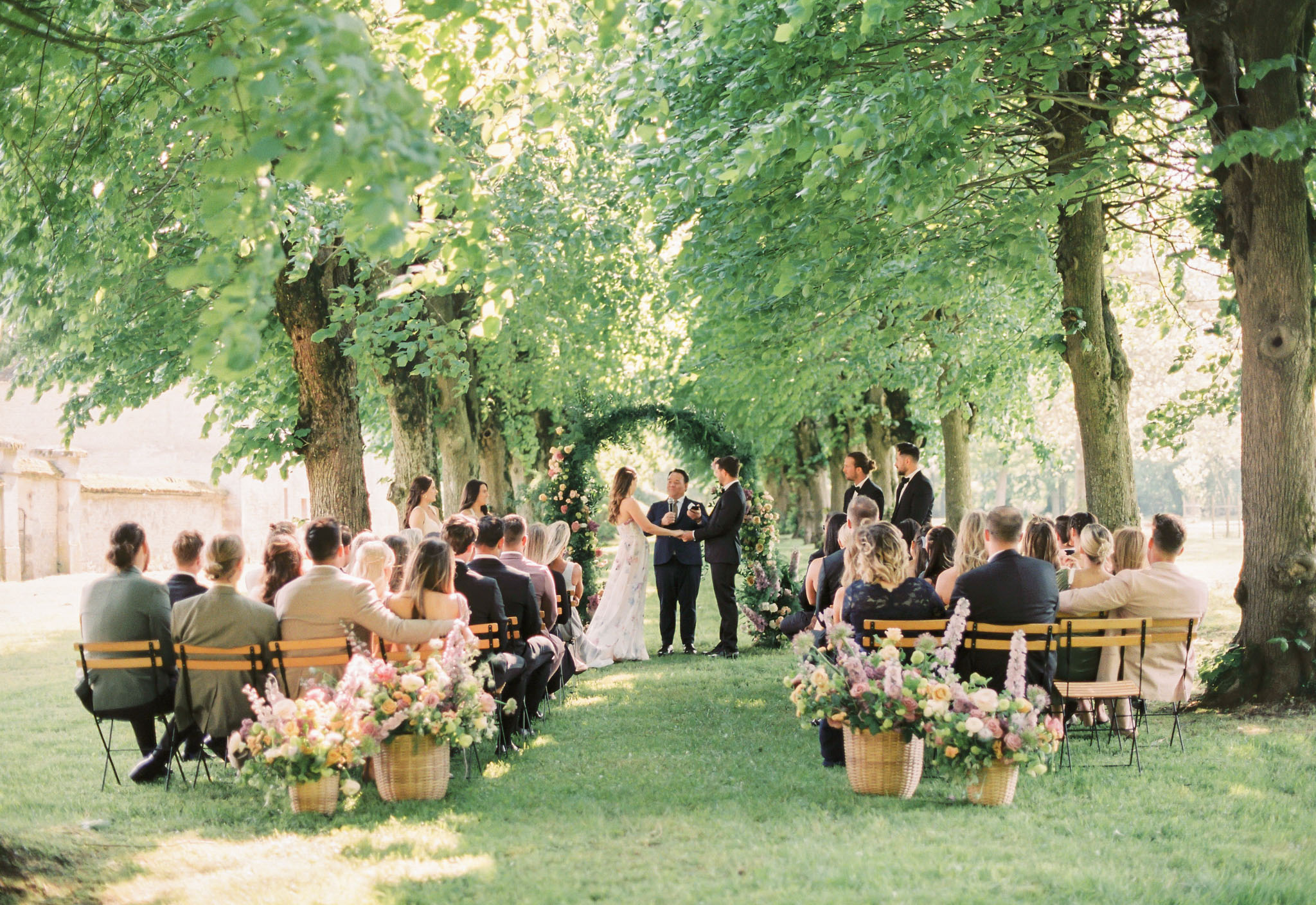 An outdoor wedding ceremony taking place along a tree-lined allée, with the couple exchanging vows at the center. The ceremony is set on a lawn beneath a canopy of mature trees, with an officiant and two groomsmen in dark navy suits standing at the altar. The bride wears a floral-printed gown in soft pastel tones, while the groom is dressed in a navy suit. Behind them stands a large circular arch covered in greenery and accented with coral, blush, and mauve florals. Approximately 40–50 guests are seated on wooden park-style benches arranged in two rows along the aisle, with wicker baskets filled with abundant mixed floral arrangements in shades of blush, peach, yellow, mauve, and coral placed at the end of each row. A bridesmaid in a champagne-toned dress stands to one side of the couple. The wide shot is taken from the rear of the ceremony space, capturing the full depth of the tree allée and a stone outbuilding partially visible to the left.