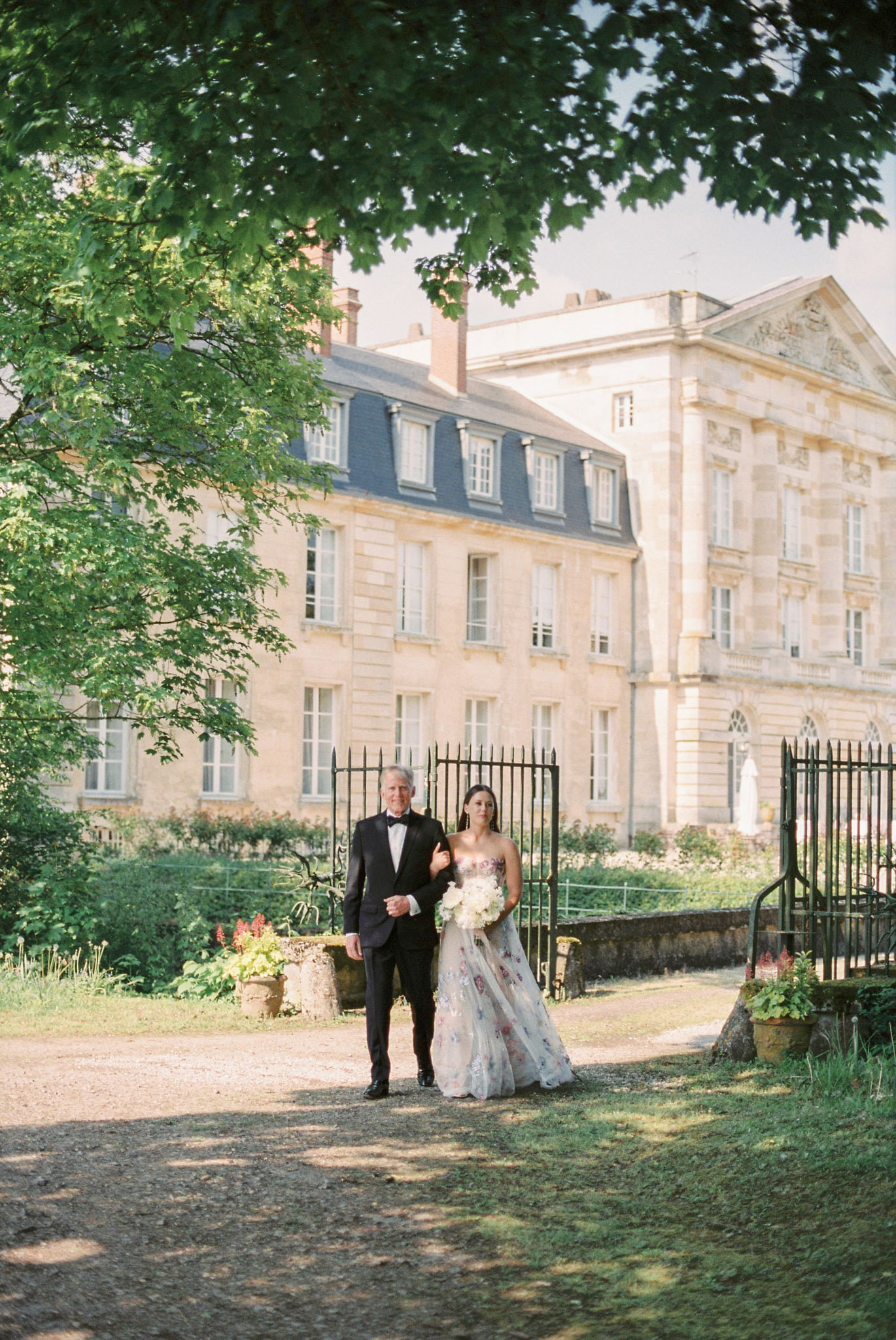 A bride is being walked down a gravel path by an older man, likely her father, during an outdoor wedding ceremony processional at a French château. The château is a large, classical French stone manor with mansard roof, dormer windows, and decorative pediment visible in the background, with ornate black iron gates flanking the path. The bride wears a strapless ball gown in ivory or pale blush with colorful floral embroidery or appliqués in pink, purple, and red tones, and carries a bouquet of white flowers including what appear to be white peonies or roses. Her escort wears a black tuxedo with bow tie. The composition is a medium-wide portrait shot with the couple centered on the path, framed by tree branches overhead and the château architecture behind them. Potential venue feature image.