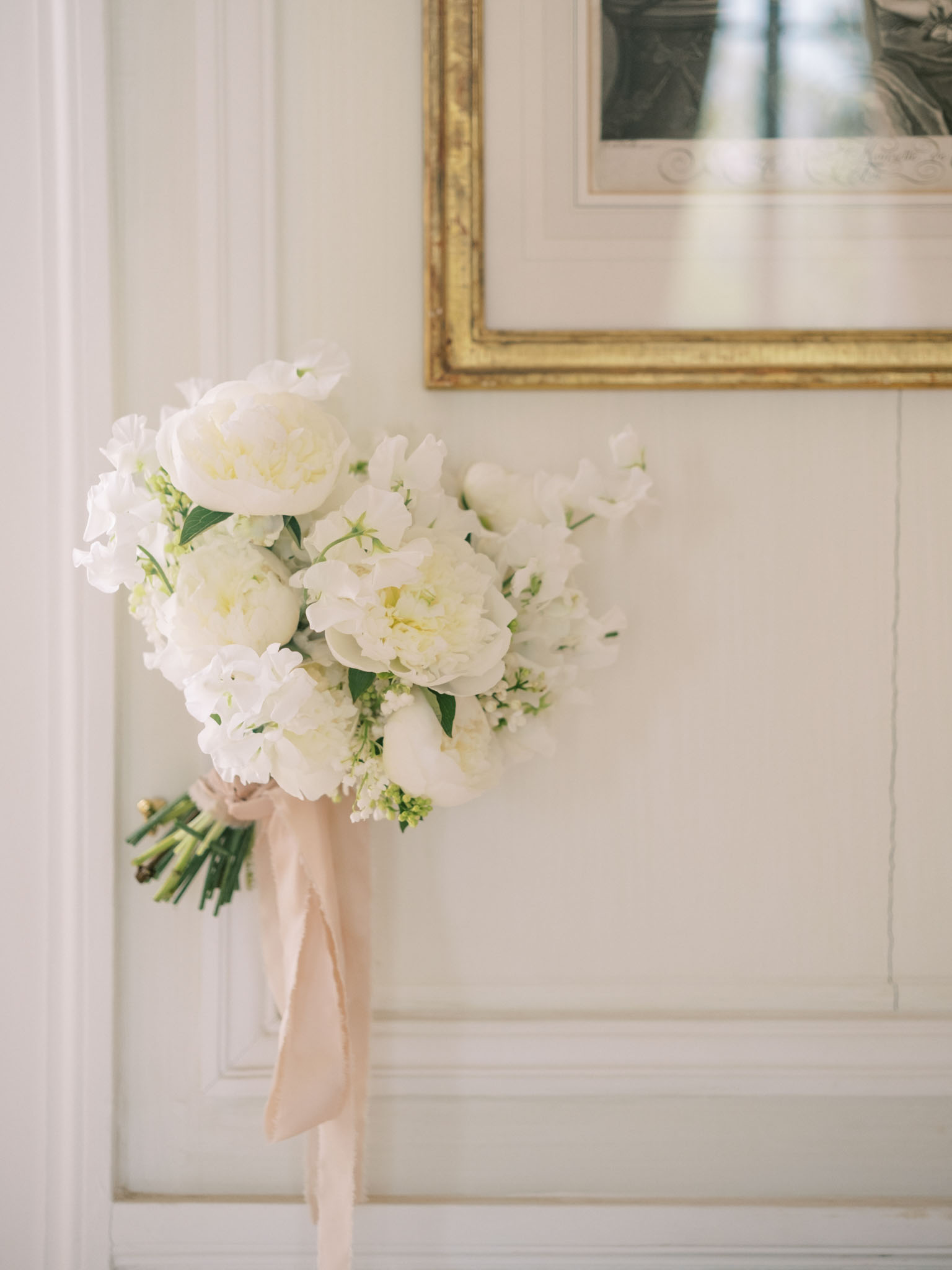 Detail shot of a bridal bouquet resting against a white paneled wall inside what appears to be a classic French interior. The bouquet is composed of white and cream peonies, white sweet peas, white hydrangea clusters, and small green filler blooms, tied with a long blush pink silk ribbon that trails downward. In the upper right corner, a gold-framed engraving or artwork is partially visible on the wall, reinforcing the classic French interior setting. The composition is a close-up still-life style shot with soft, even natural light highlighting the white and cream tones of the florals against the pale wall.