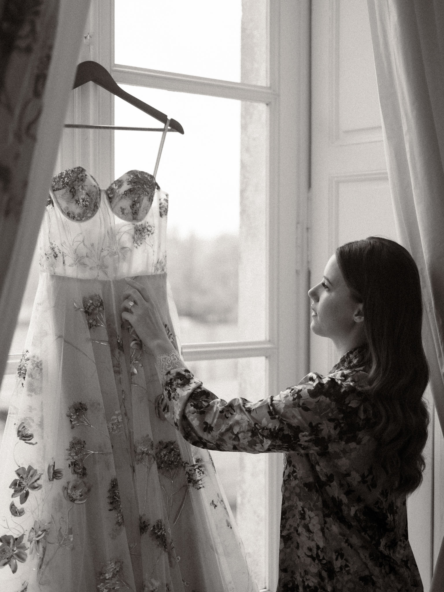 Bride reaching for embroidered tulle gown on hanger by backlit window in chateau room in B&W