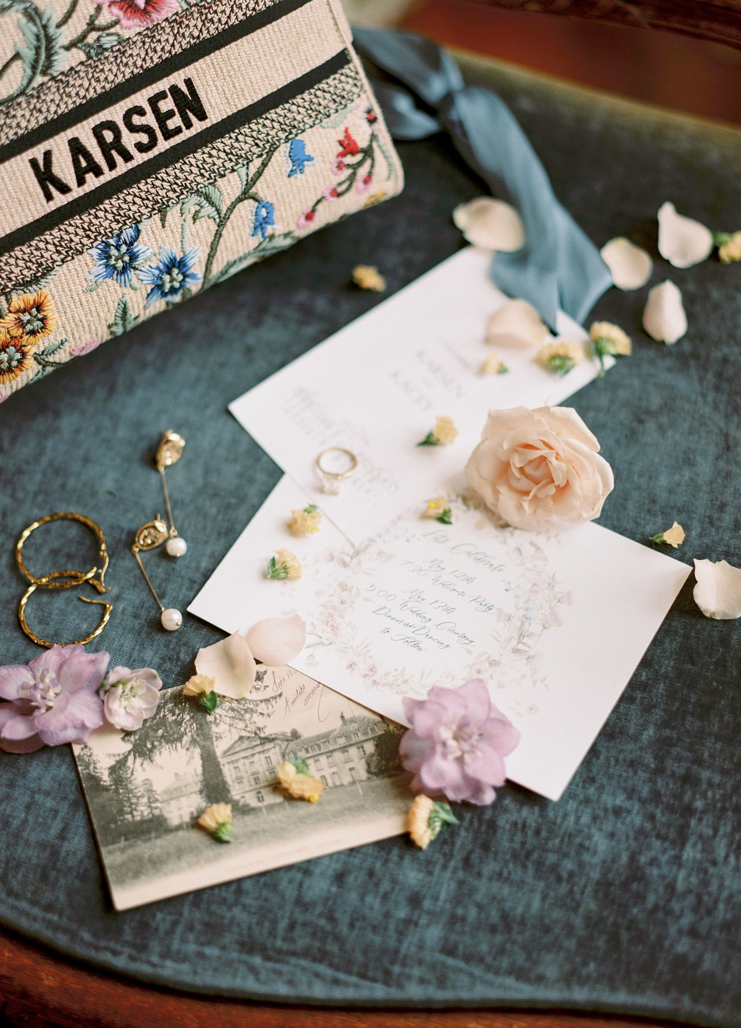 Flat lay on teal velvet with Dior tote invitation suite gold rings pearl earrings rose petals and blue ribbon