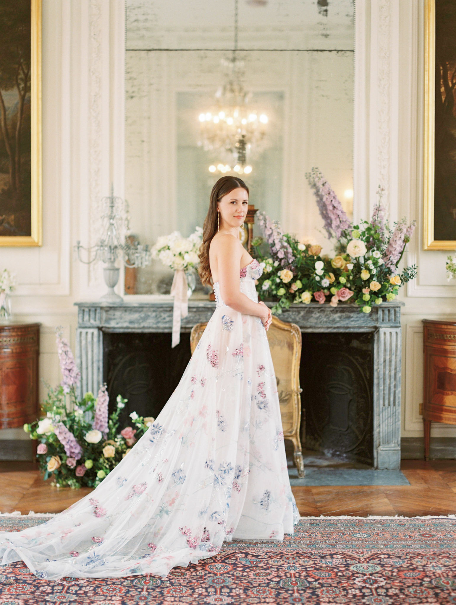 Bride in floral-embroidered strapless gown looking over shoulder before ornate marble fireplace with garden flower arrange...