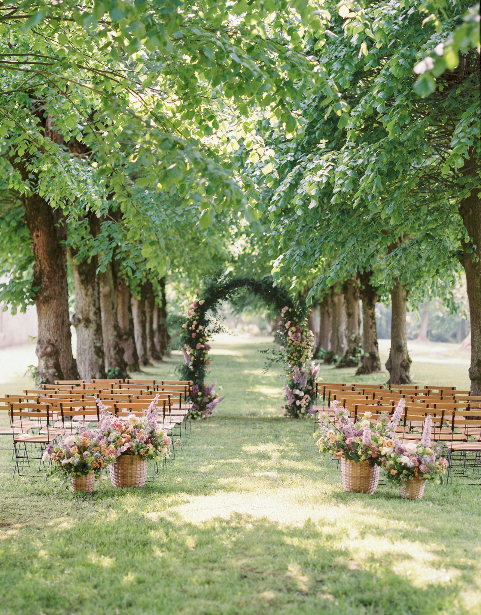 Tree-lined ceremony aisle with peach and mauve floral arch and wicker basket arrangements for 100 guests