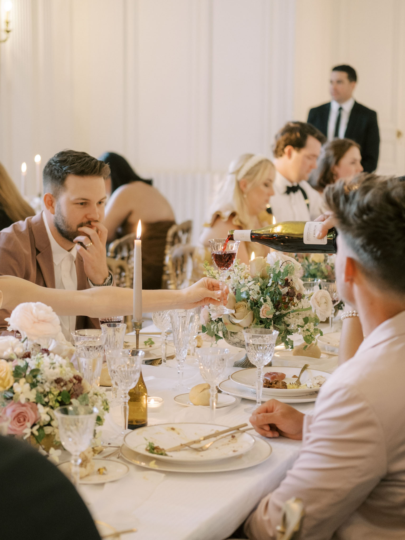 An indoor wedding reception dinner is underway in a formal white-panelled ballroom, with a server pouring red wine from a bottle into a crystal glass at a long banquet table. Approximately six to eight guests are visible seated along the table, with a man in a dusty rose blazer prominently in the foreground and a woman in a white dress and pearl bracelet seated across from him. The table is set with white linens, gold-rimmed china plates, crystal glassware, and gold flatware, with lit taper candles in brass holders and low floral centerpieces featuring ivory garden roses, blush and mauve roses, burgundy accents, and trailing greenery. In the background, a man in a black tuxedo with a bow tie and a woman in a white dress — likely the couple — are seated further down the table, while a staff member in a dark suit stands nearby. The overall decor palette is ivory, blush, gold, and soft green, consistent with a classic French formal reception setting. The shot is taken from a medium distance at table level, with shallow depth of field creating a candid, editorial feel.