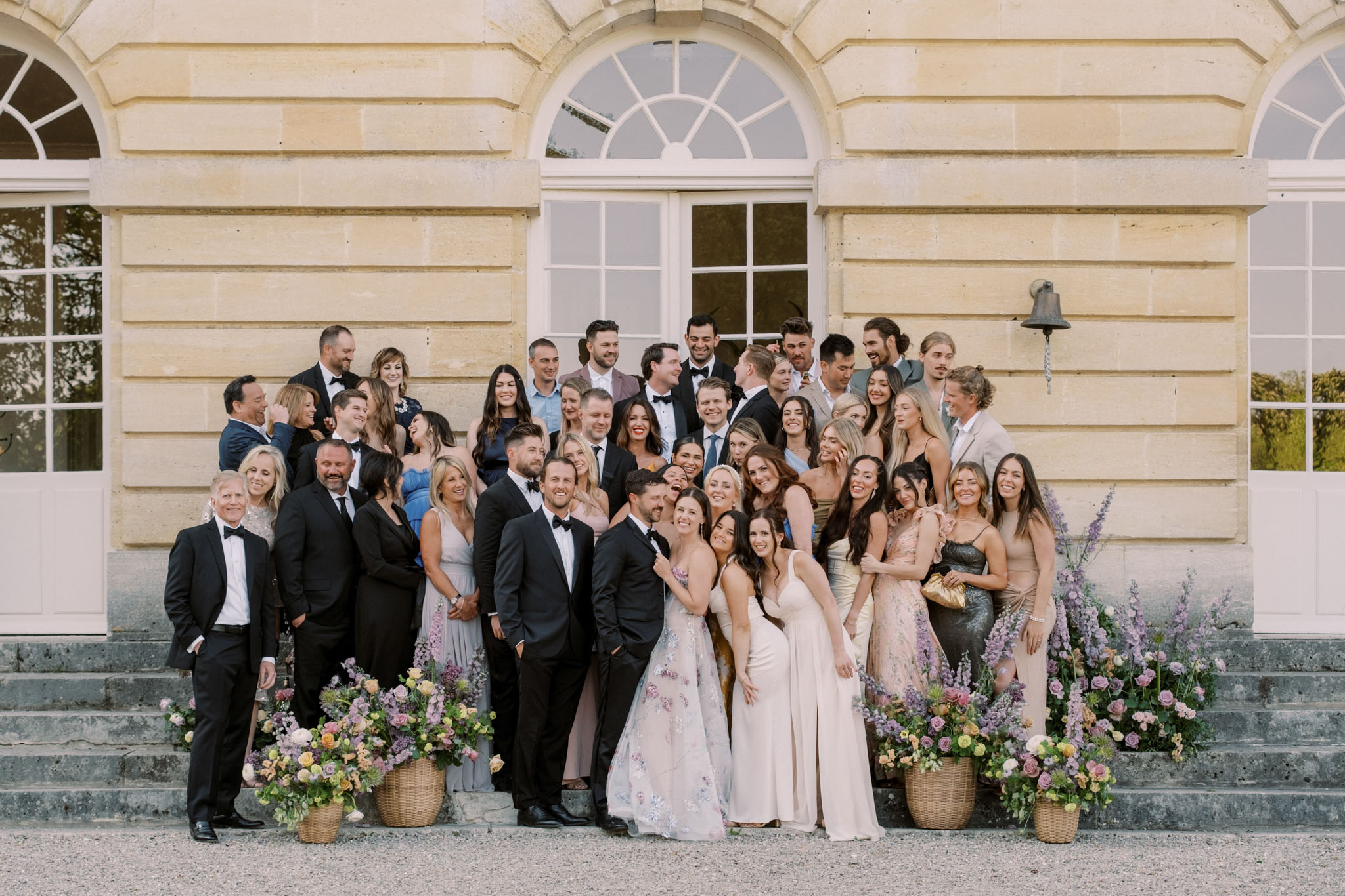 Wedding group portrait of 40 guests on neoclassical chateau steps flanked by lavender and blush floral baskets