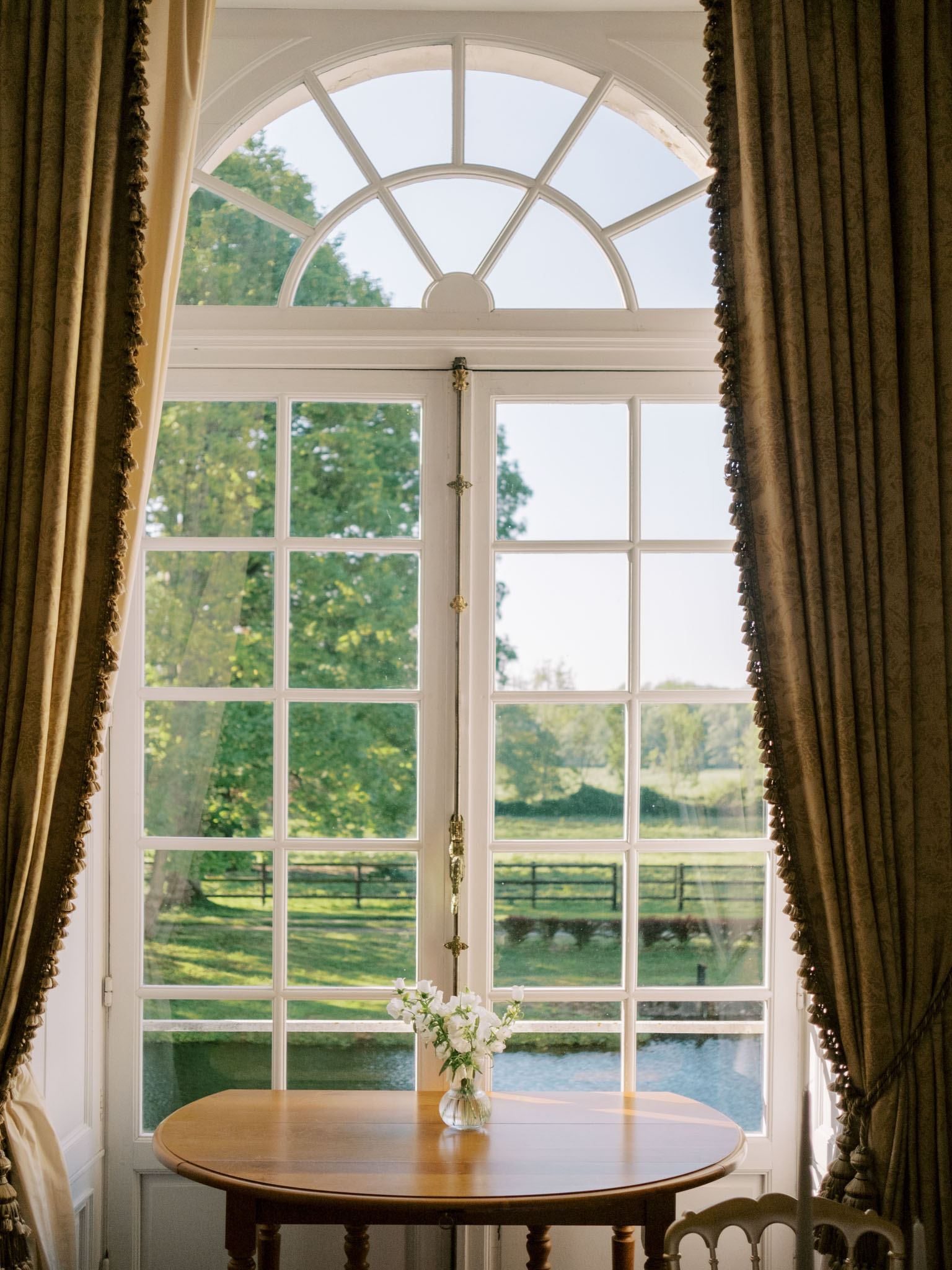 Arched chateau window with olive velvet curtains and white sweet peas in glass vase on round table