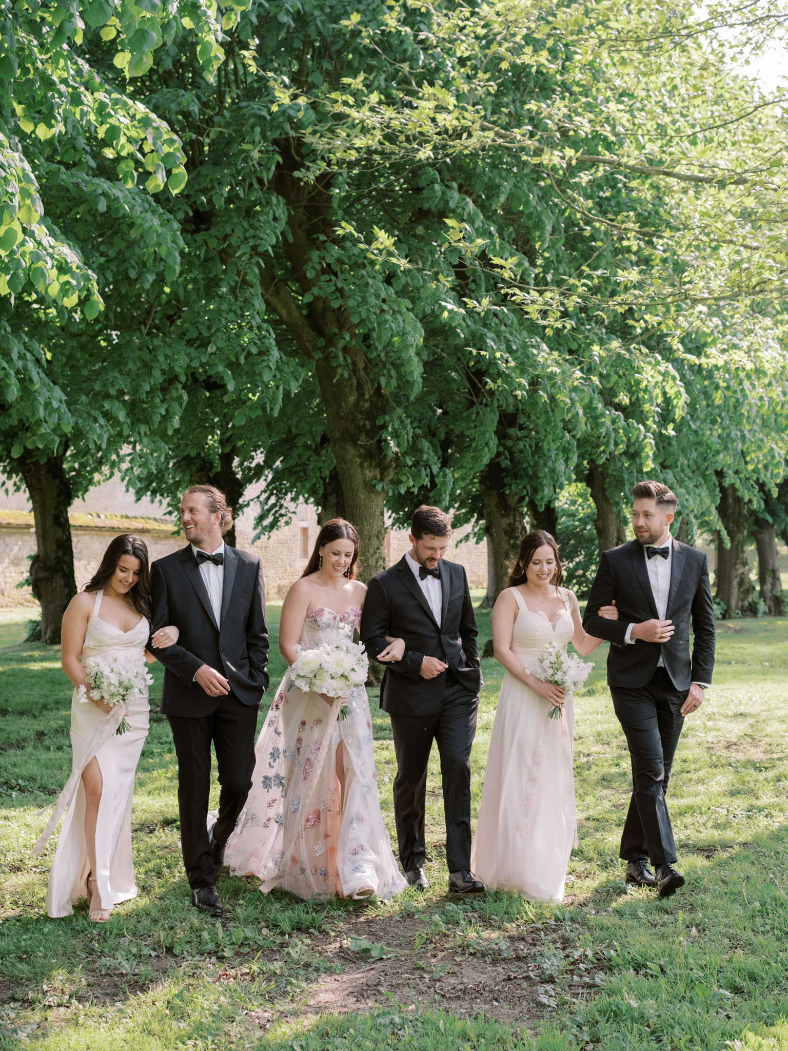 Bridal party of six walking along tree-lined avenue with bride in floral applique gown and blush bridesmaids