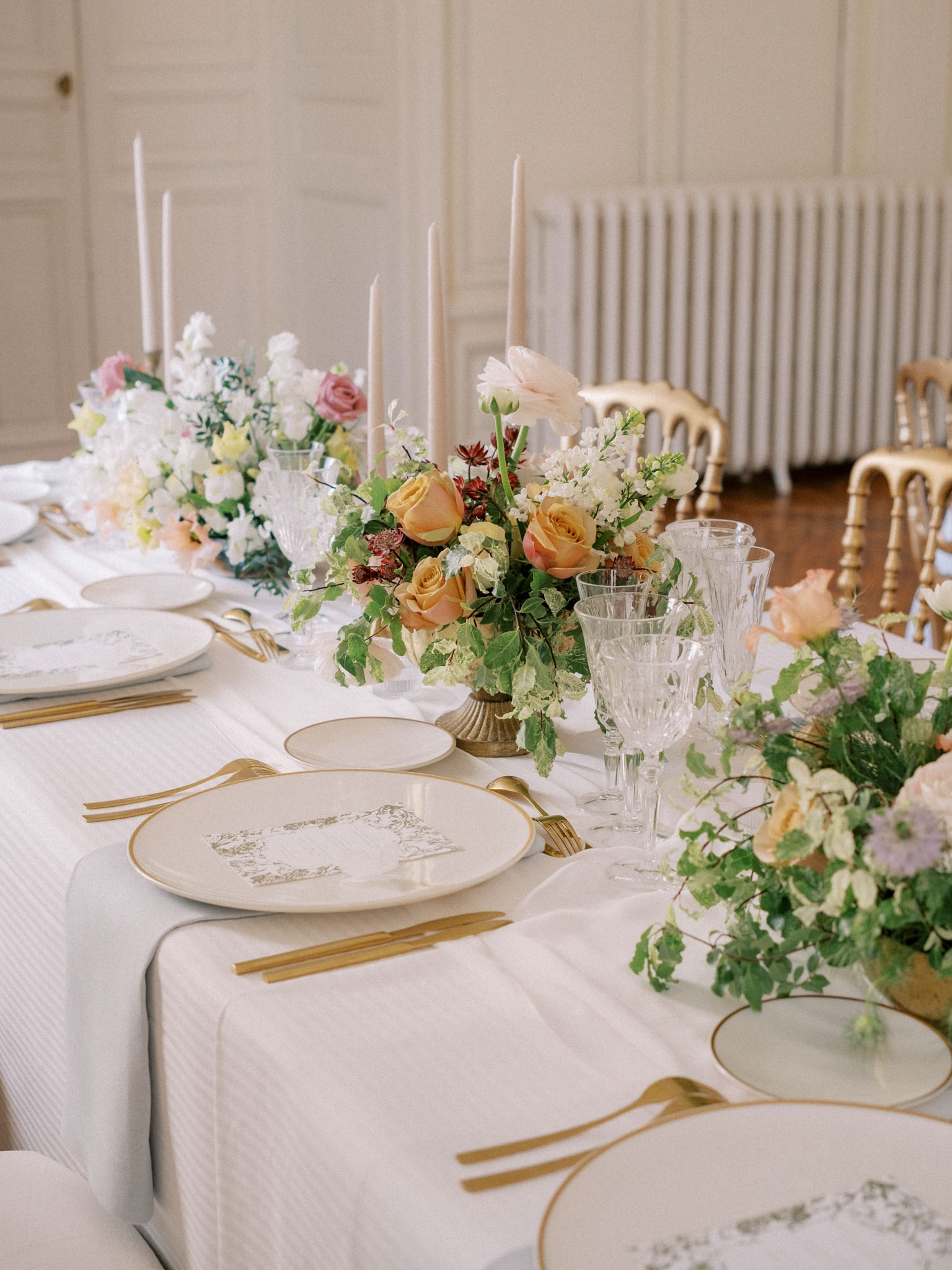 Wedding reception table with gold-rimmed chargers, amber and peach rose centerpieces in brass urns, and blush taper candles