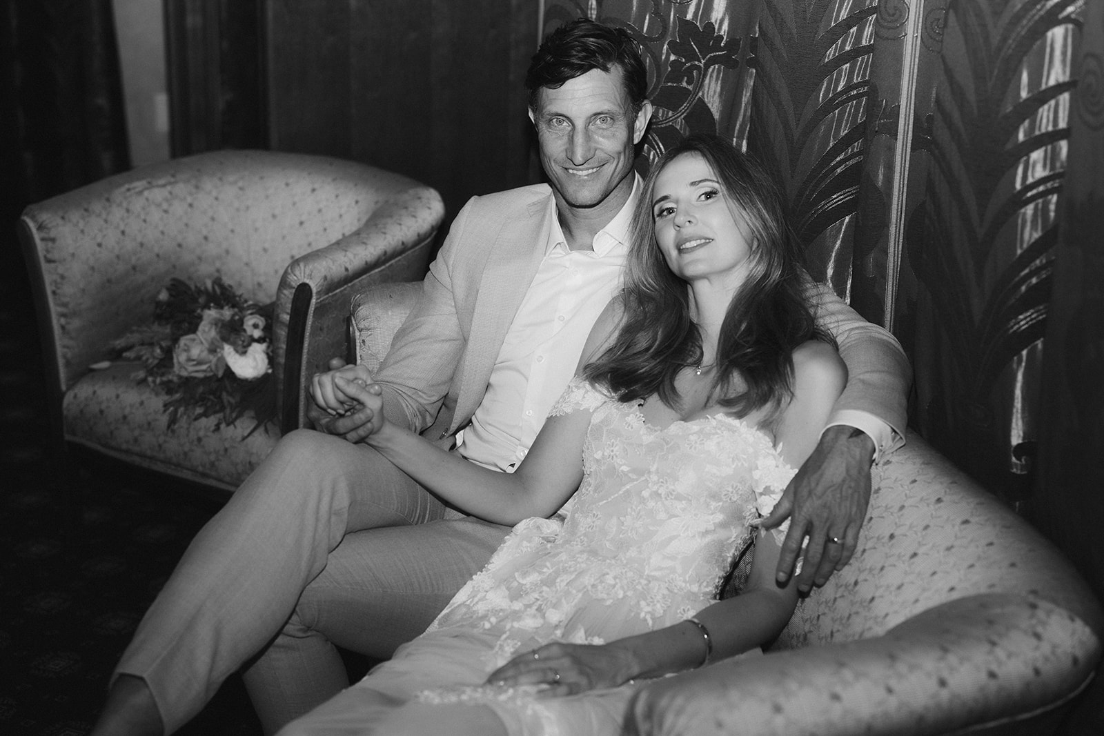 Black and white couple relaxing on sofa bride in lace gown groom in blazer with botanical mural behind
