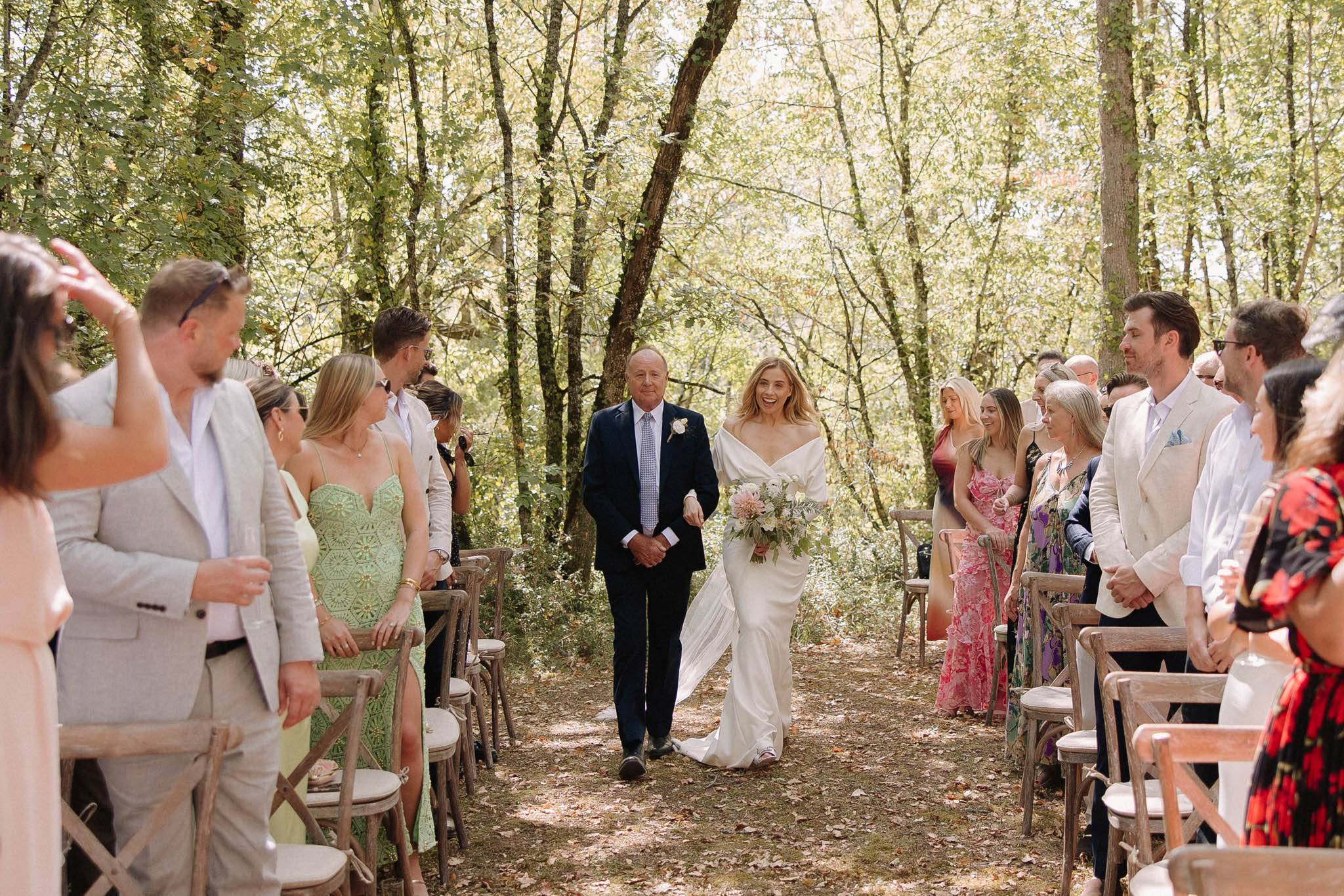 Bride walked down woodland aisle by father during outdoor forest ceremony with rustic cross-back chairs