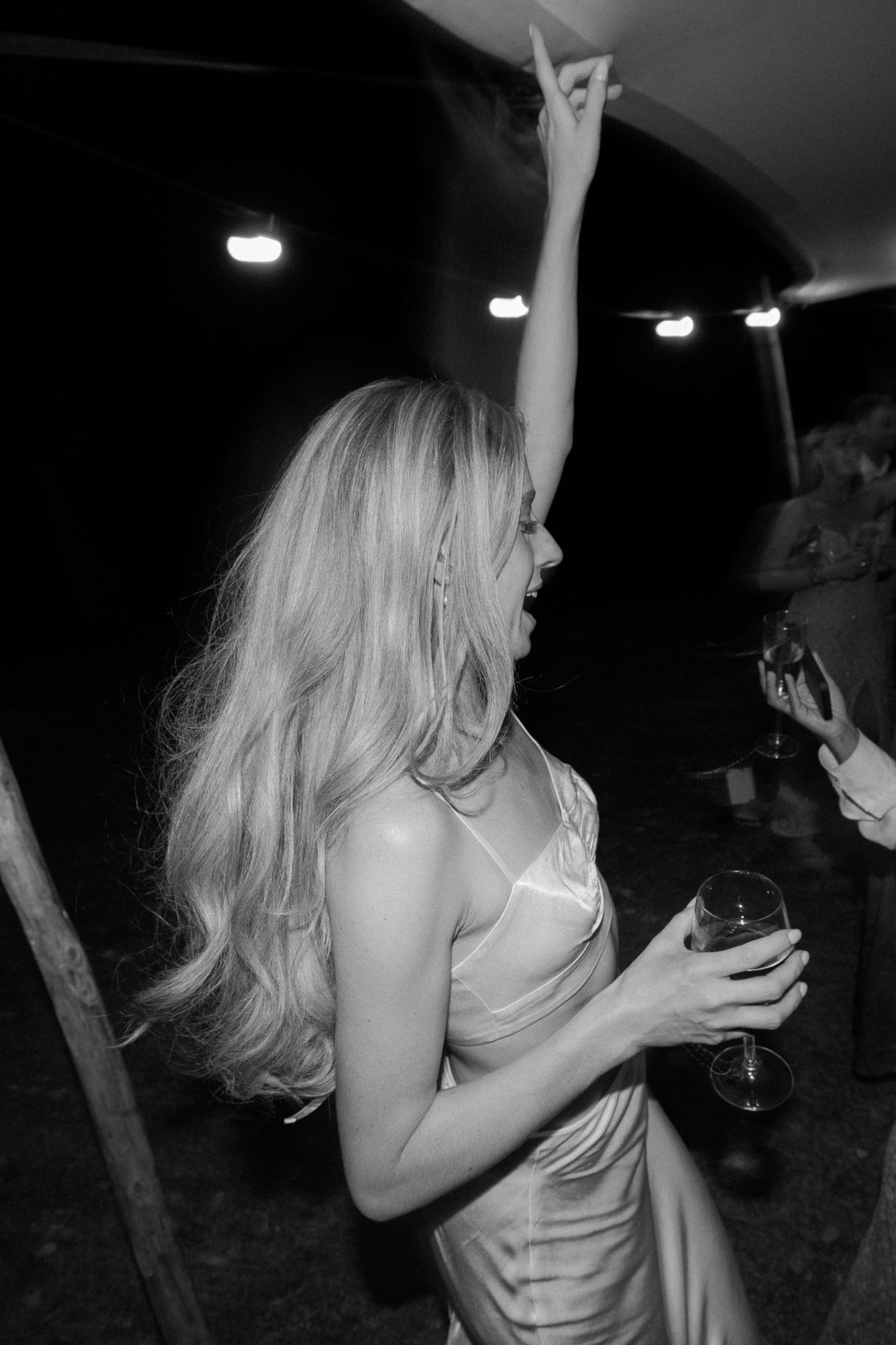 A black-and-white candid shot taken during an evening wedding reception, likely inside a marquee or tented venue, with overhead spot lighting visible in the dark background. A woman with long, loose wavy hair is dancing with one arm raised, holding a wine glass in her other hand, laughing and turned slightly away from the camera. She is wearing a satin slip-style dress with thin straps, consistent with a satin bridesmaid or guest dress. At least two other guests are partially visible in the background, one holding a stemmed glass. The high-contrast black-and-white tones give the image a grainy, documentary feel, with deep blacks in the background and bright highlights on the subject's hair and dress. The composition is a close portrait shot from a low, slightly angled perspective, capturing movement and energy on the dance floor.