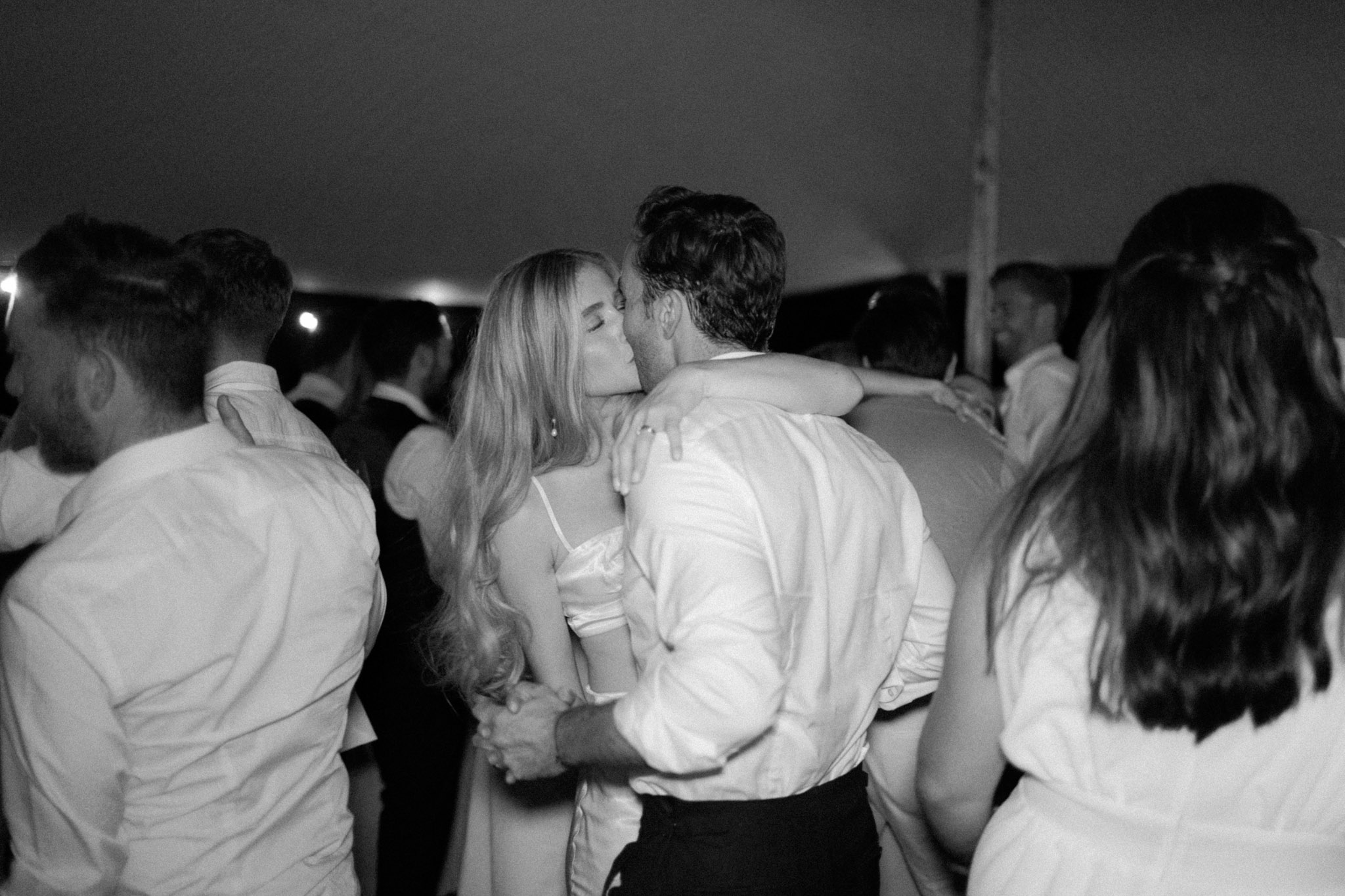 Black-and-white candid of couple kissing on crowded dance floor during tent reception