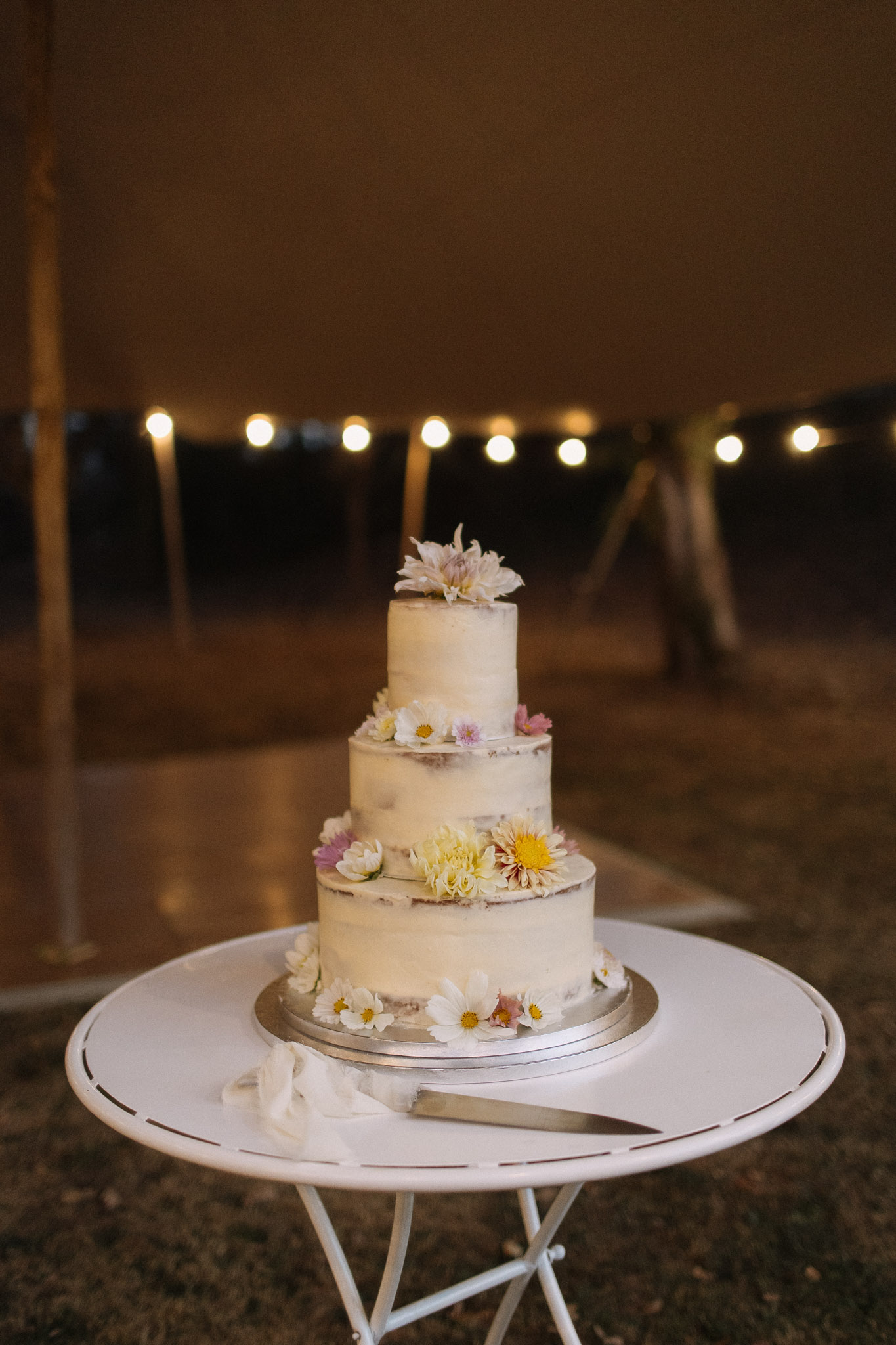 Three-tier semi-naked cake with fresh cosmos and dahlias on bistro table inside tipi tent
