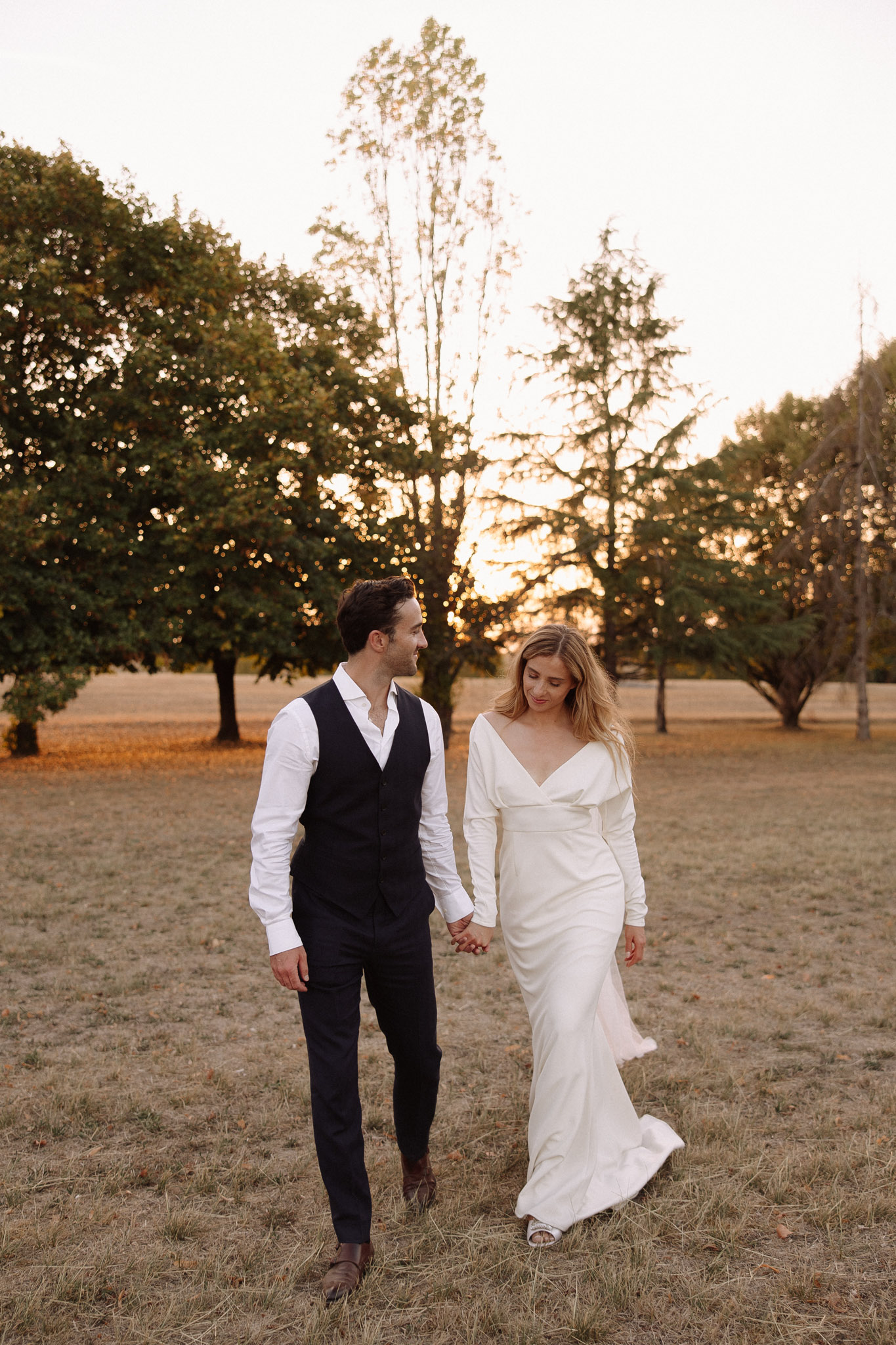 A couple portrait taken outdoors in an open field during golden hour, with warm backlight filtering through a tree line in the background. The bride wears a long-sleeve ivory satin slip-style gown with a deep V-neckline and a fitted silhouette, paired with open-toe heeled sandals, while the groom wears dark trousers, a black waistcoat, and an open-collar white dress shirt with brown leather shoes. The two are walking hand in hand across dry grass, the groom looking toward the bride and the bride looking downward. The overall styling is modern and minimal, with a clean, understated aesthetic. Medium full-length portrait shot.