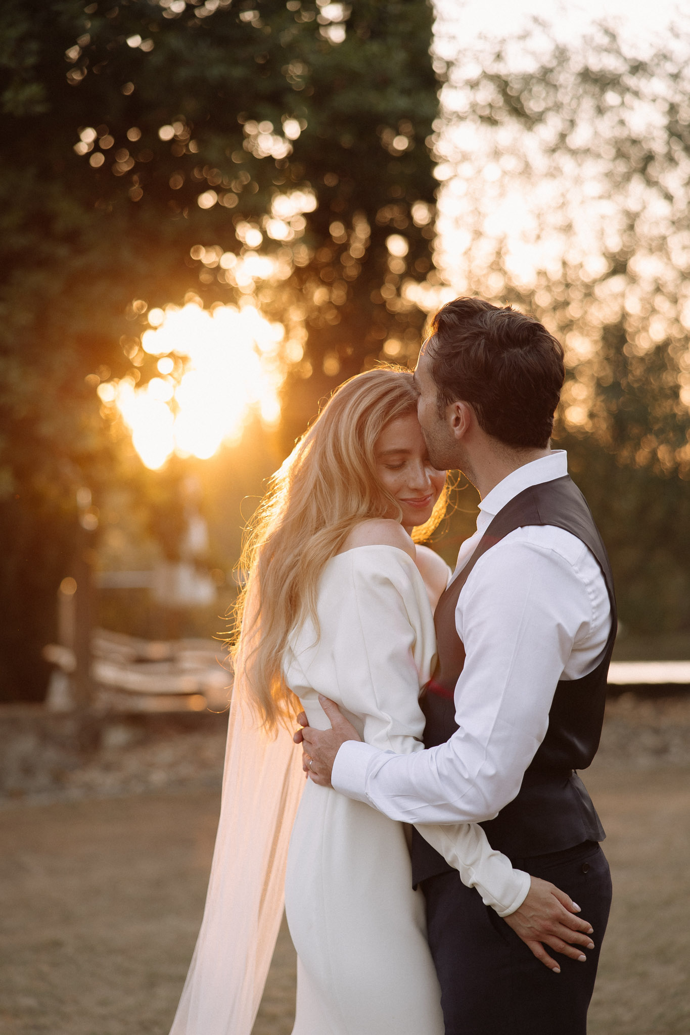 A couple portrait taken outdoors during golden hour, with warm sunset light filtering through trees in the background creating strong bokeh. The bride has long blonde hair and wears an off-the-shoulder ivory dress with long sleeves and a flowing cape or veil train; the groom wears a white dress shirt with a dark charcoal waistcoat and dark trousers, his jacket removed. The groom leans in to kiss the bride's temple as she smiles with her eyes closed, the two embracing in a relaxed, candid pose. The composition is a medium portrait shot with the backlit sunset used as a dramatic light source, giving the image a warm amber and gold tone.