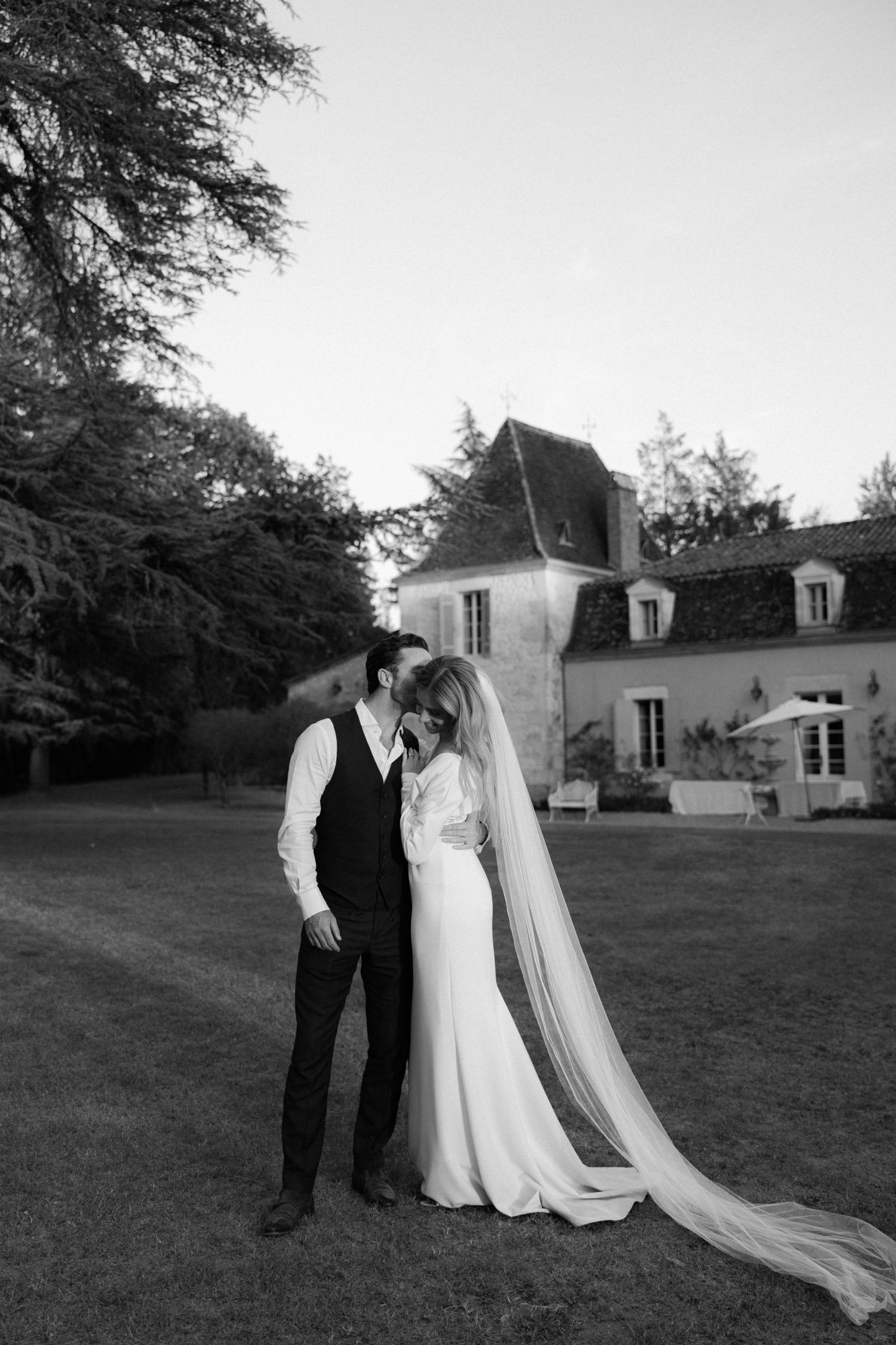 A black-and-white couple portrait taken outdoors on the grounds of a French château, with the manor house and its steep-pitched roof tower visible in the background. The groom, dressed in a dark suit with a waistcoat and open-collar white shirt, leans in to kiss the bride on the cheek while embracing her from behind. The bride wears a long-sleeved, slim-fitting white gown with a cathedral-length veil that trails across the lawn. The image has strong mid-tone contrast with bright highlights on the bride's dress and veil against the darker tones of the grounds and trees. The composition is a full-length portrait shot with the château centered in the background, anchoring the couple within the estate setting.