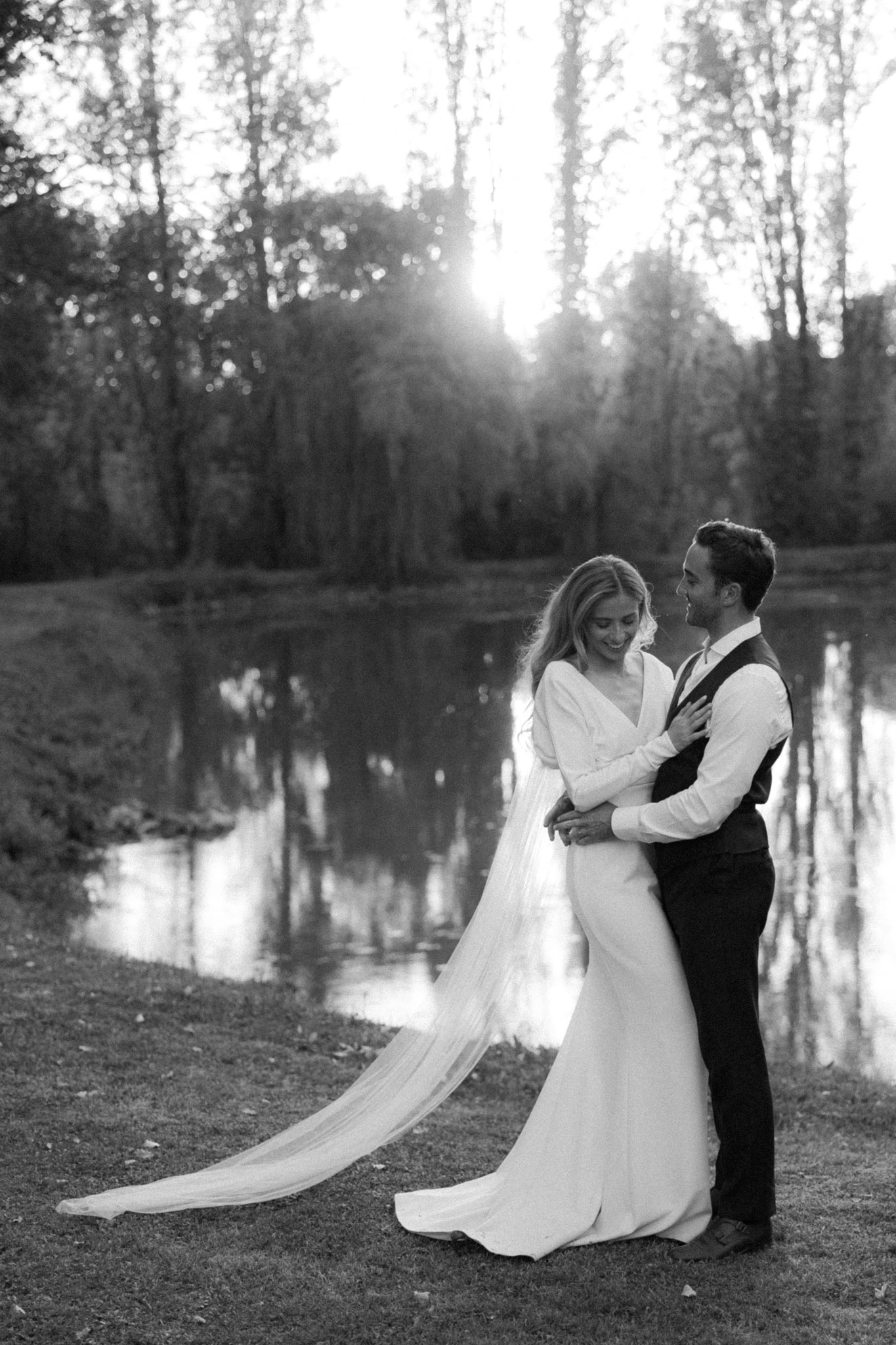 Black-and-white couple portrait embracing beside a pond with cathedral-length veil and backlit trees