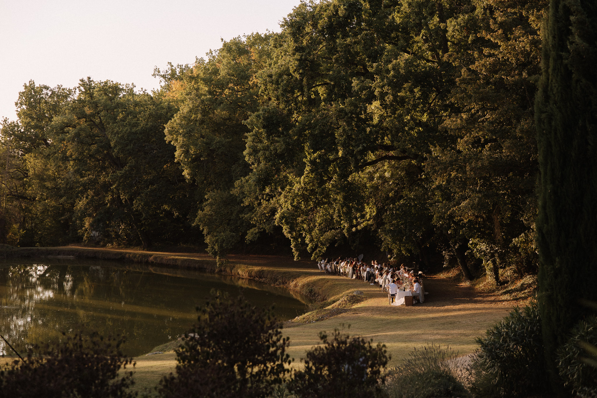 An outdoor wedding reception dinner is taking place along the bank of a calm ornamental lake, with approximately 40–60 guests seated at what appears to be a single long table running parallel to the water's edge. The setting is a large private estate or château grounds, with the table positioned on a flat lawn strip between the lake and a dense line of mature oak trees. Warm golden-hour light bathes the scene, casting long shadows across the grass. Table linens appear white or ivory, and floral centerpieces in warm, earthy tones are faintly visible at the near end of the table. The overall styling reads as classic and natural, making use of the landscape as the primary decorative element. This is a wide establishing shot taken from an elevated vantage point, showing the full length of the dining setup within its landscape context. Potential venue feature image.