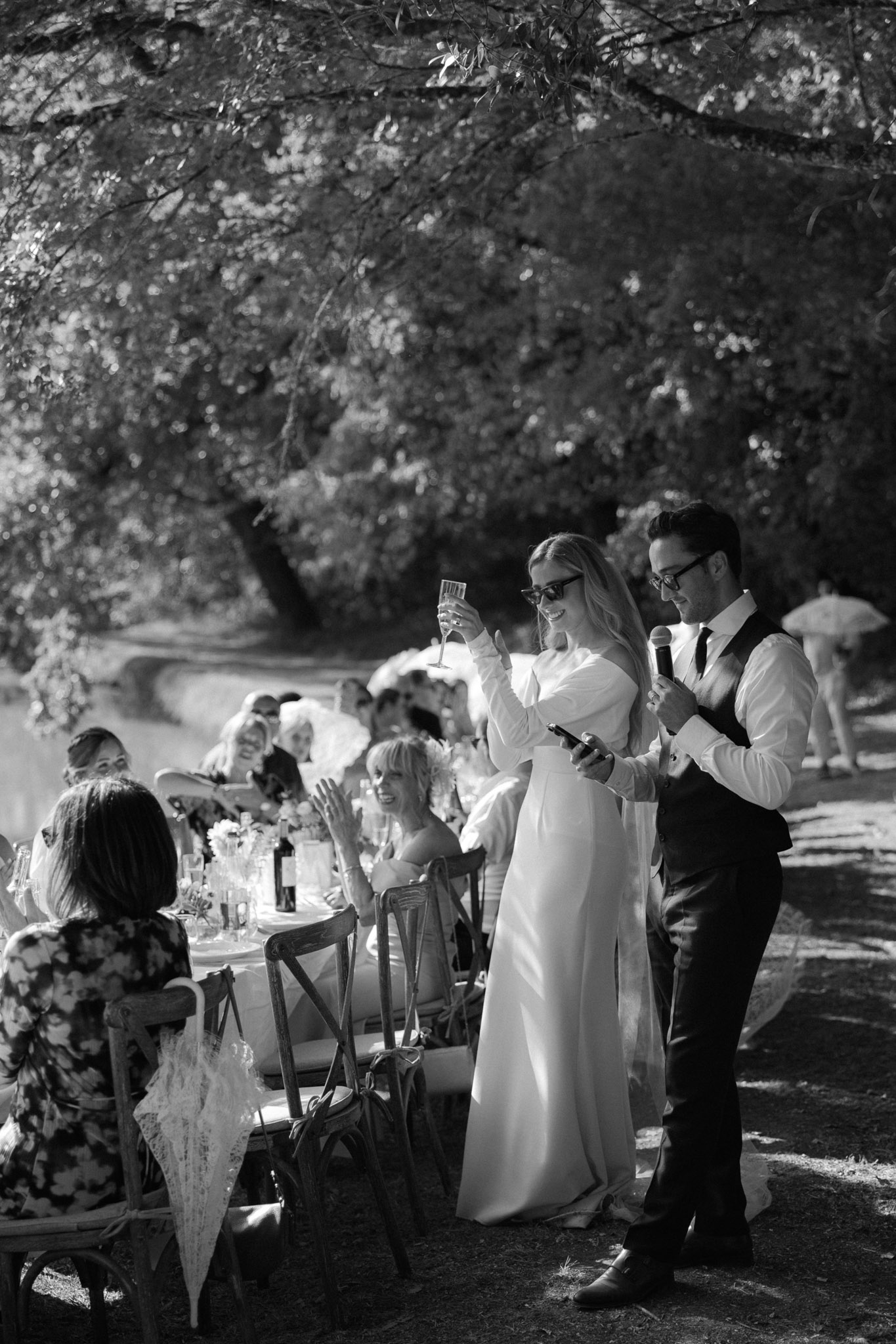 Black and white image of bride and groom giving speech at outdoor reception with guests at round tables