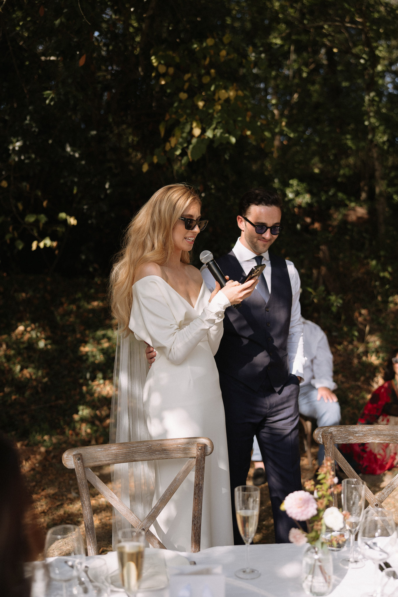 Bride and groom giving speech together wearing sunglasses at outdoor reception table with pink and red flowers