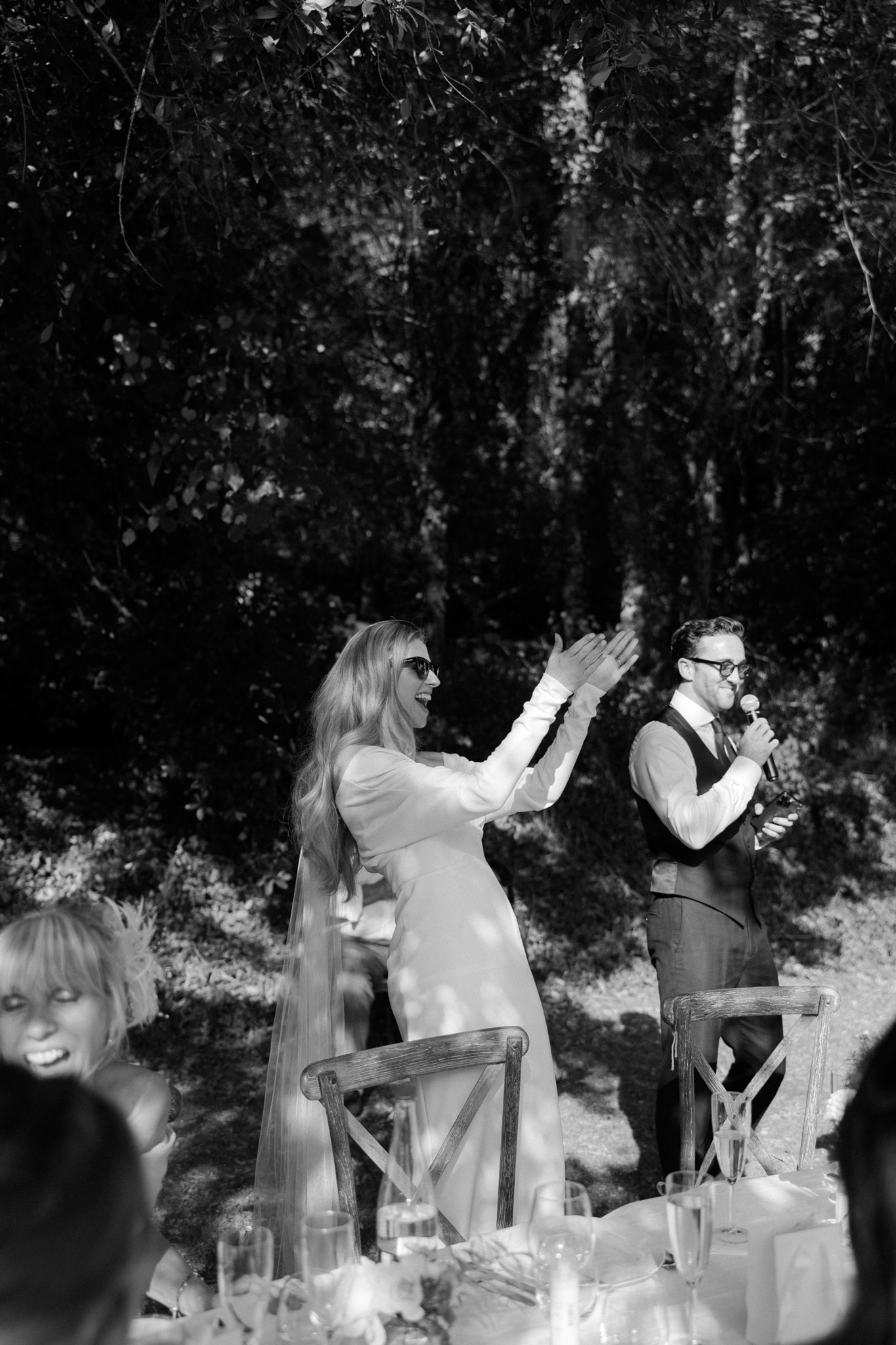 A black-and-white image capturing a speech moment during an outdoor wedding reception, set against a backdrop of dense trees with dappled sunlight. The bride, wearing a long-sleeved white dress and a long veil with sunglasses, is standing and clapping enthusiastically with a wide smile, while the groom stands beside her in a dark waistcoat and trousers, holding a microphone and a bottle. A female guest in the foreground, partially visible, is also laughing. The reception table in the lower portion of the frame is set with champagne flutes, candles, and floral arrangements, with rustic cross-back wooden chairs visible. The high contrast black-and-white tones give the scene a bright, sun-drenched quality with deep shadows in the tree canopy above.