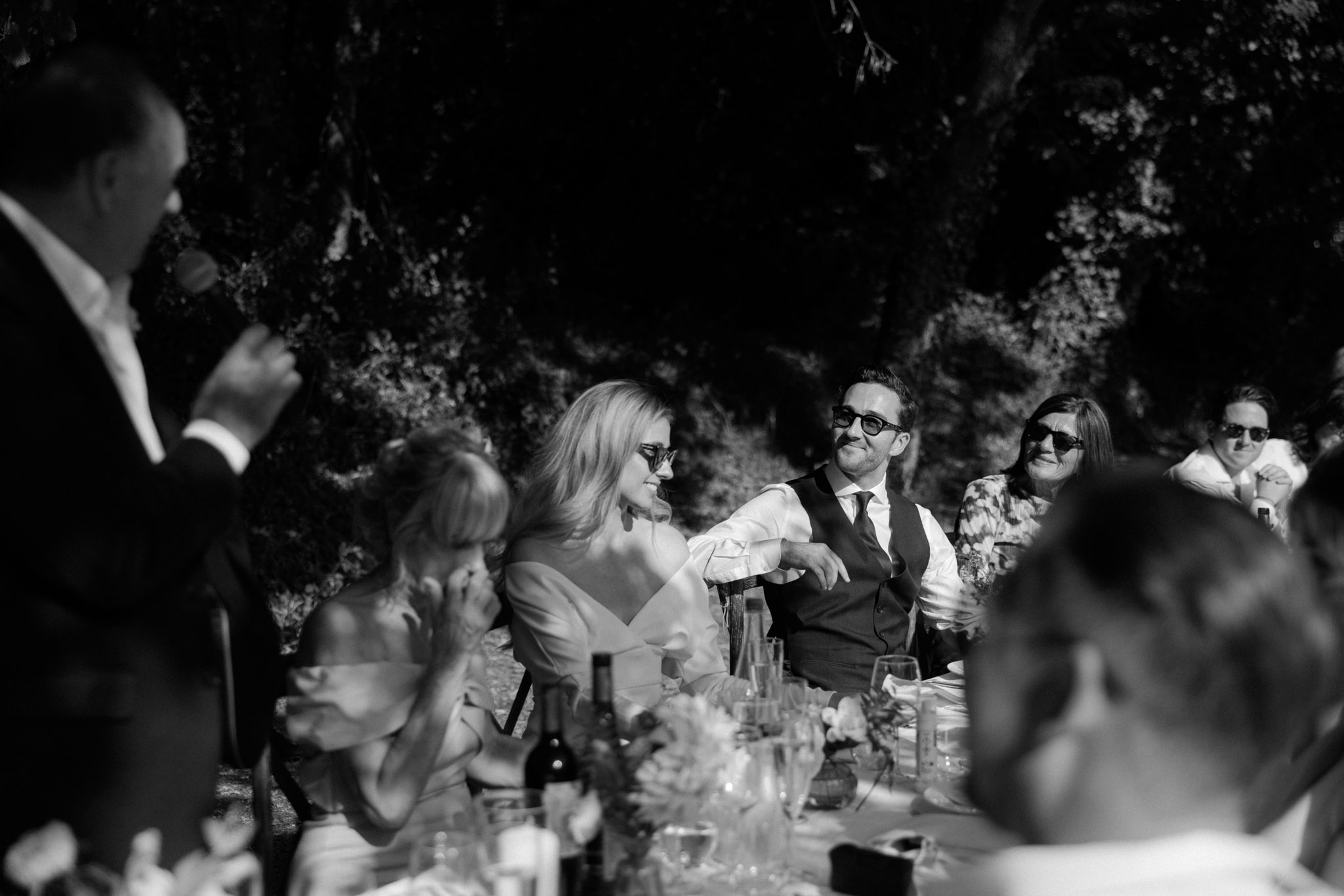 Black-and-white photo of bride and groom reacting to speech at outdoor reception table in sunlight