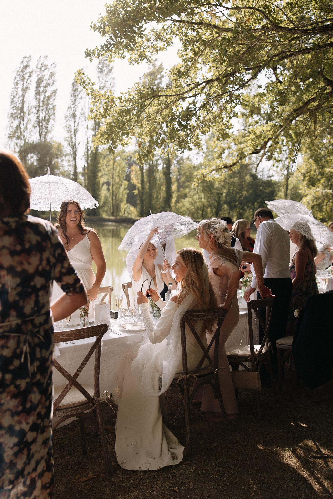 An outdoor wedding reception meal is taking place beside a lake or river, with approximately 10–15 guests visible gathered around a long table dressed in a white linen cloth. The bride, wearing a long-sleeved ivory fitted gown, is seated at the table in a rustic wooden cross-back chair, holding sunglasses and looking upward while conversing with guests. Several guests are holding white lace parasols to shade themselves from the bright afternoon sun. A bridesmaid or guest in a short white dress stands nearby laughing. Table settings include champagne flutes, small bud vases with greenery, and white napkins. Seating consists of wooden cross-back chairs throughout. The overall styling is relaxed and rustic-boho, with the lace parasols as a coordinated decorative detail. The shot is a medium-wide candid portrait taken at a slight angle, capturing the lively atmosphere of the outdoor dining setting.