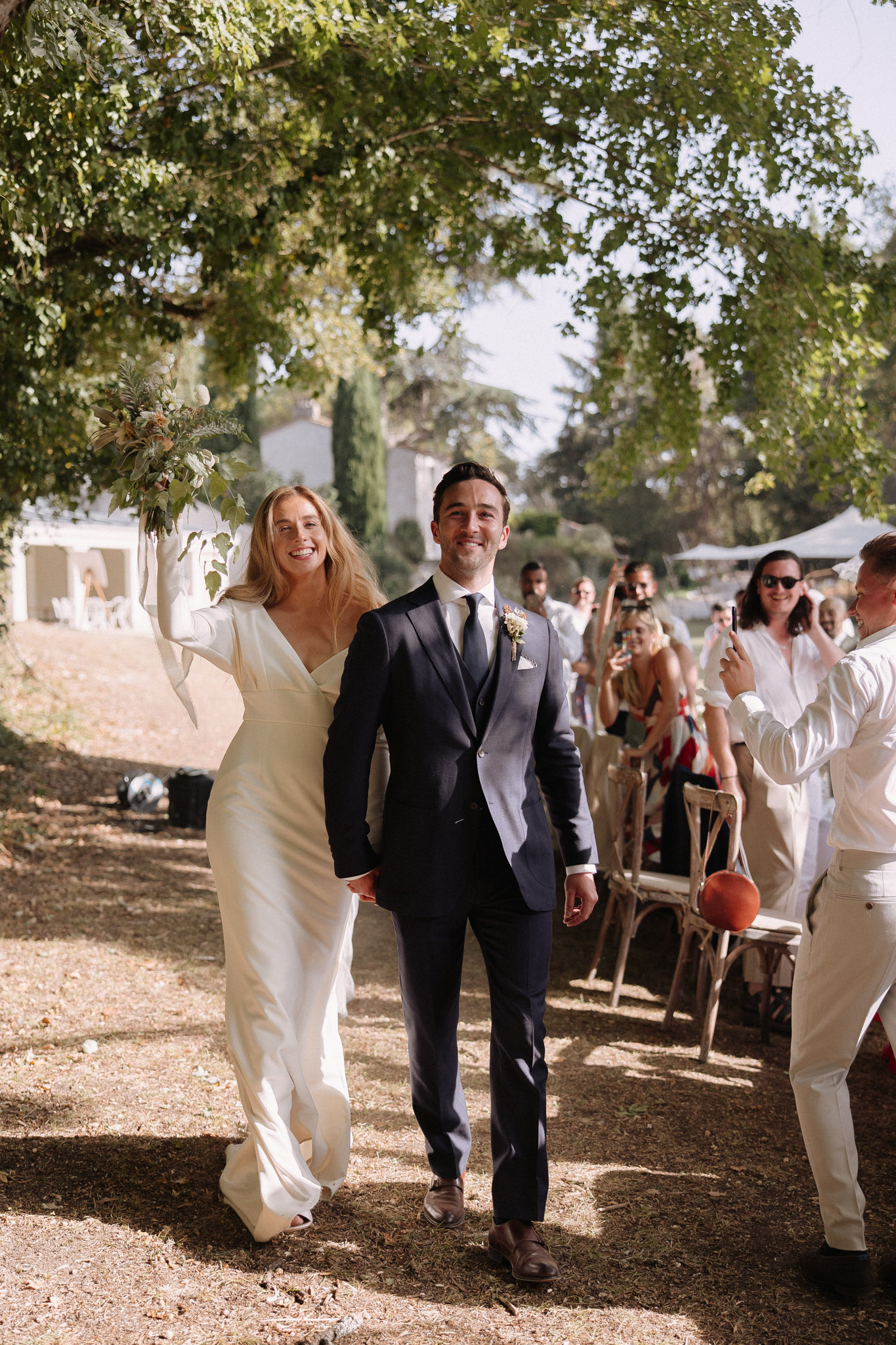 Bride and groom recessioning as guests cheer with bride raising greenery bouquet at country manor