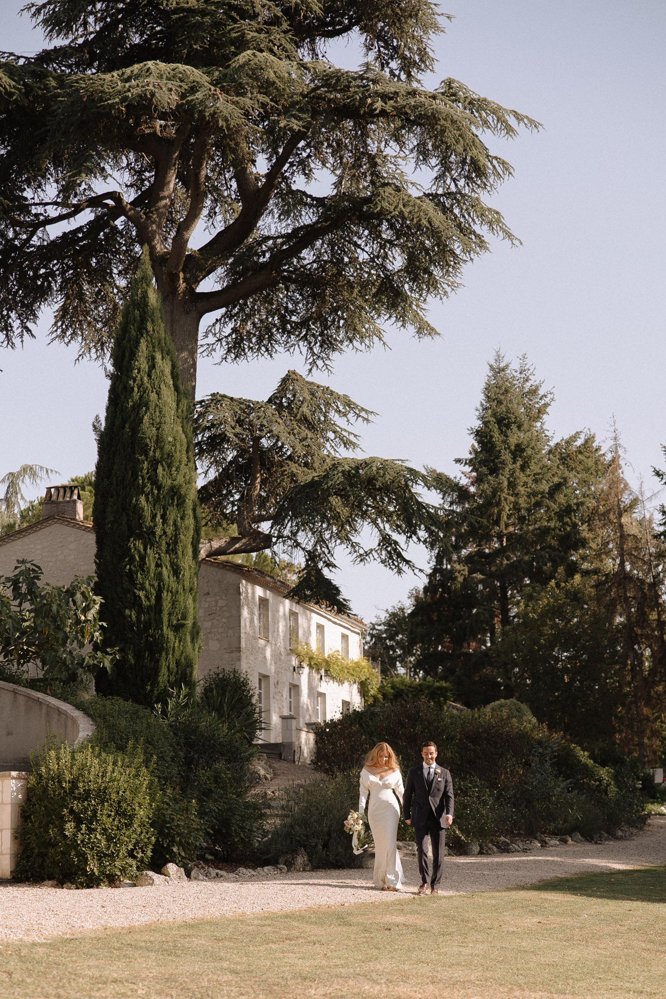 Bride and groom walking hand in hand on gravel path with stone manor house, cedar tree, and cypress behind