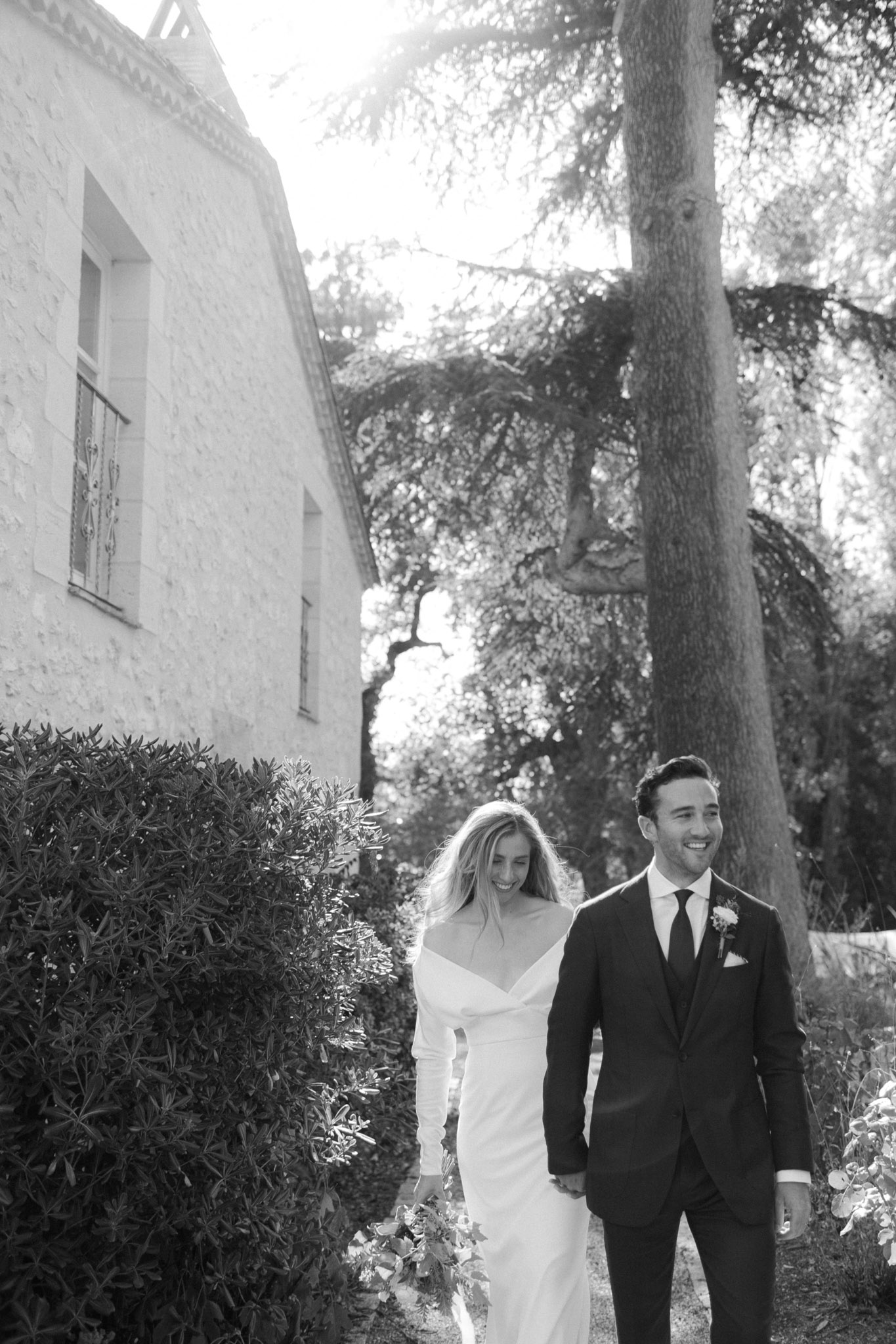 Black-and-white portrait of bride and groom walking and laughing hand in hand on chateau grounds with backlit trees