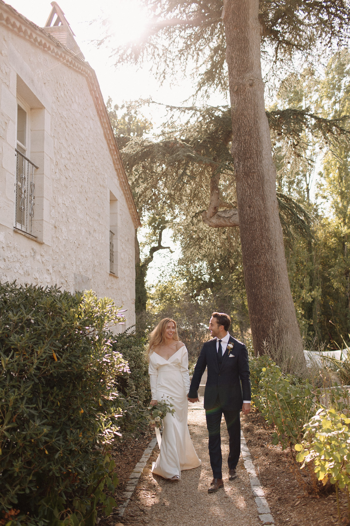Couple walks hand-in-hand along limestone manor path with golden backlit sun flare through trees