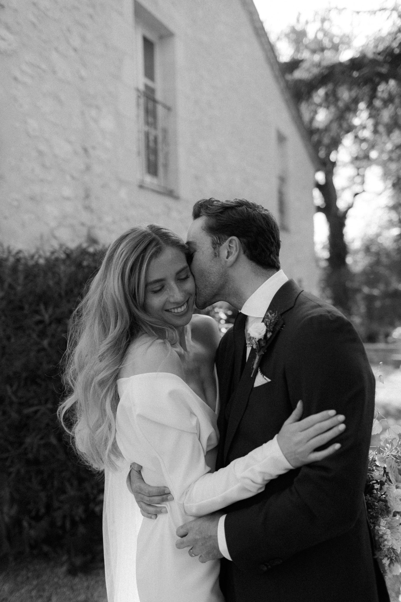 This is a black-and-white couple portrait taken outdoors against the whitewashed stone facade of what appears to be a French country property or château. The groom, dressed in a dark suit with a tie and a floral boutonnière featuring a light-toned bloom, leans in to kiss the bride on the temple. The bride wears an off-the-shoulder gown with long sleeves and long white gloves, and her wavy blonde hair falls loosely over her shoulders; she is smiling with her eyes closed. The two are embracing, with the groom's hands around her waist and her arms wrapped around him. The image has soft, even contrast with bright highlights on the bride's dress and gentle mid-tones throughout. The composition is a close-up portrait with the building and trimmed hedging softly out of focus in the background.