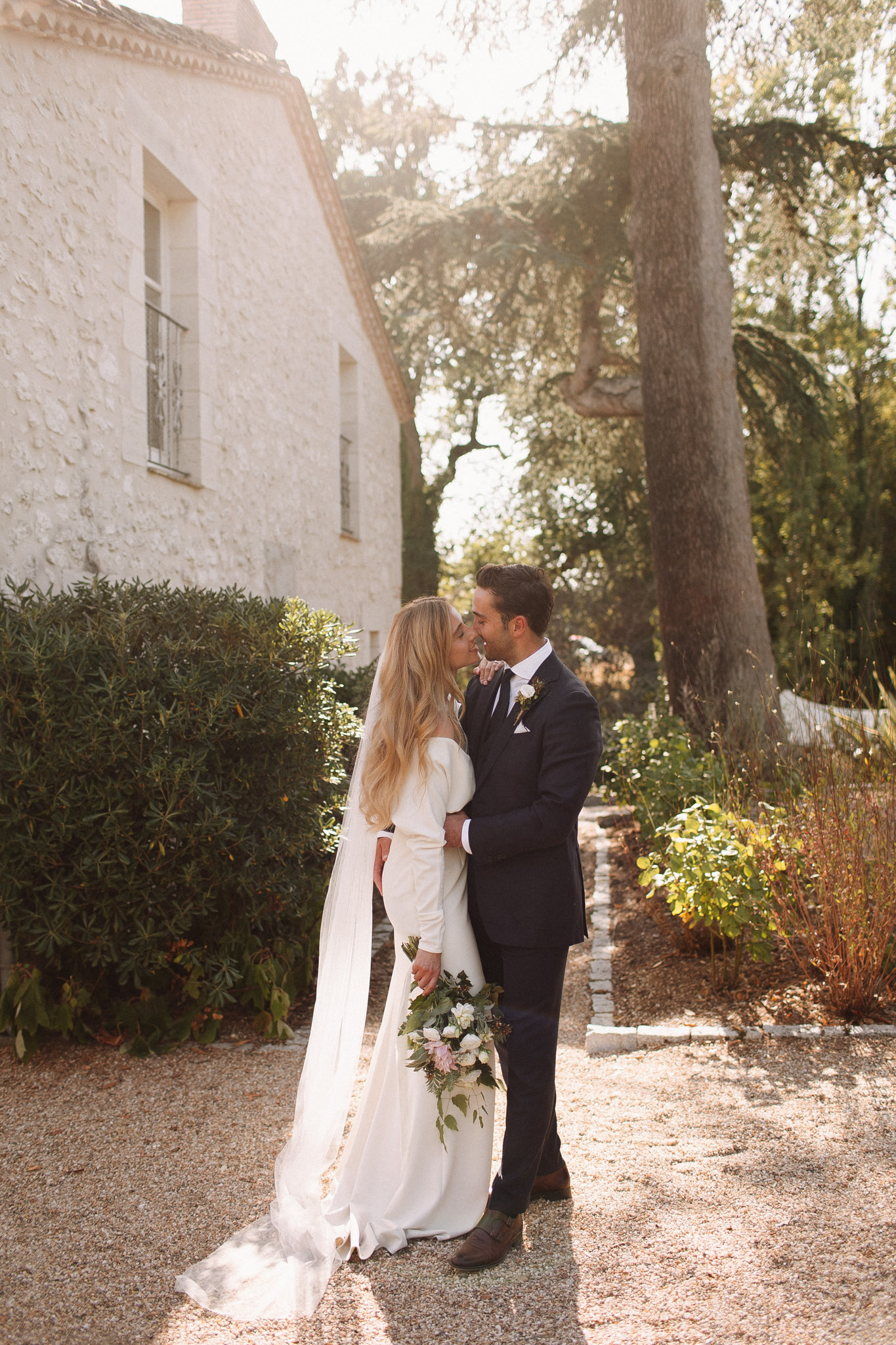 Bride and groom kissing on a gravel path beside a stone building with golden backlight through trees