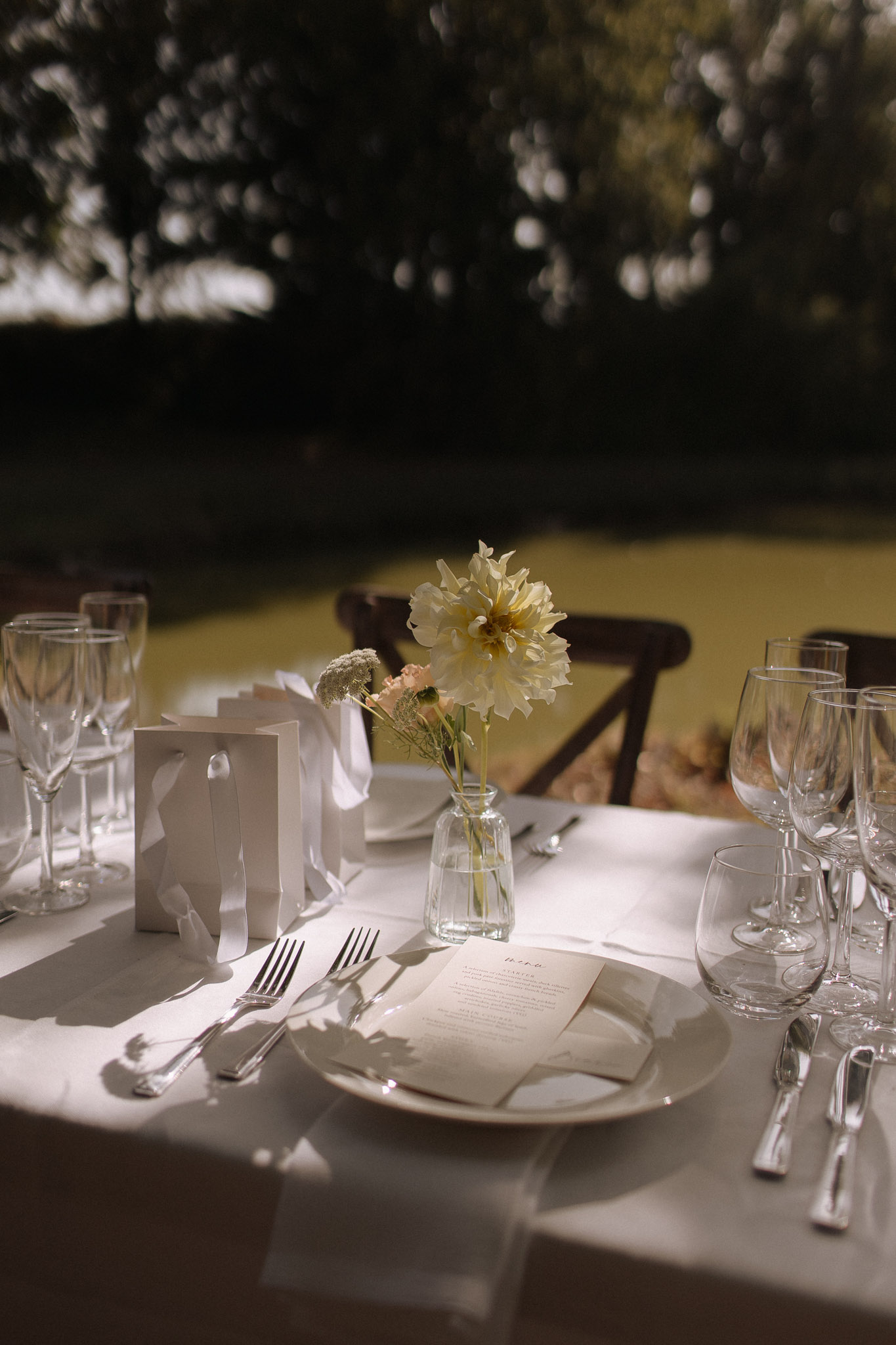 Reception table place setting with white plates, crystal glasses, cream dahlia bud vase, and printed menu card