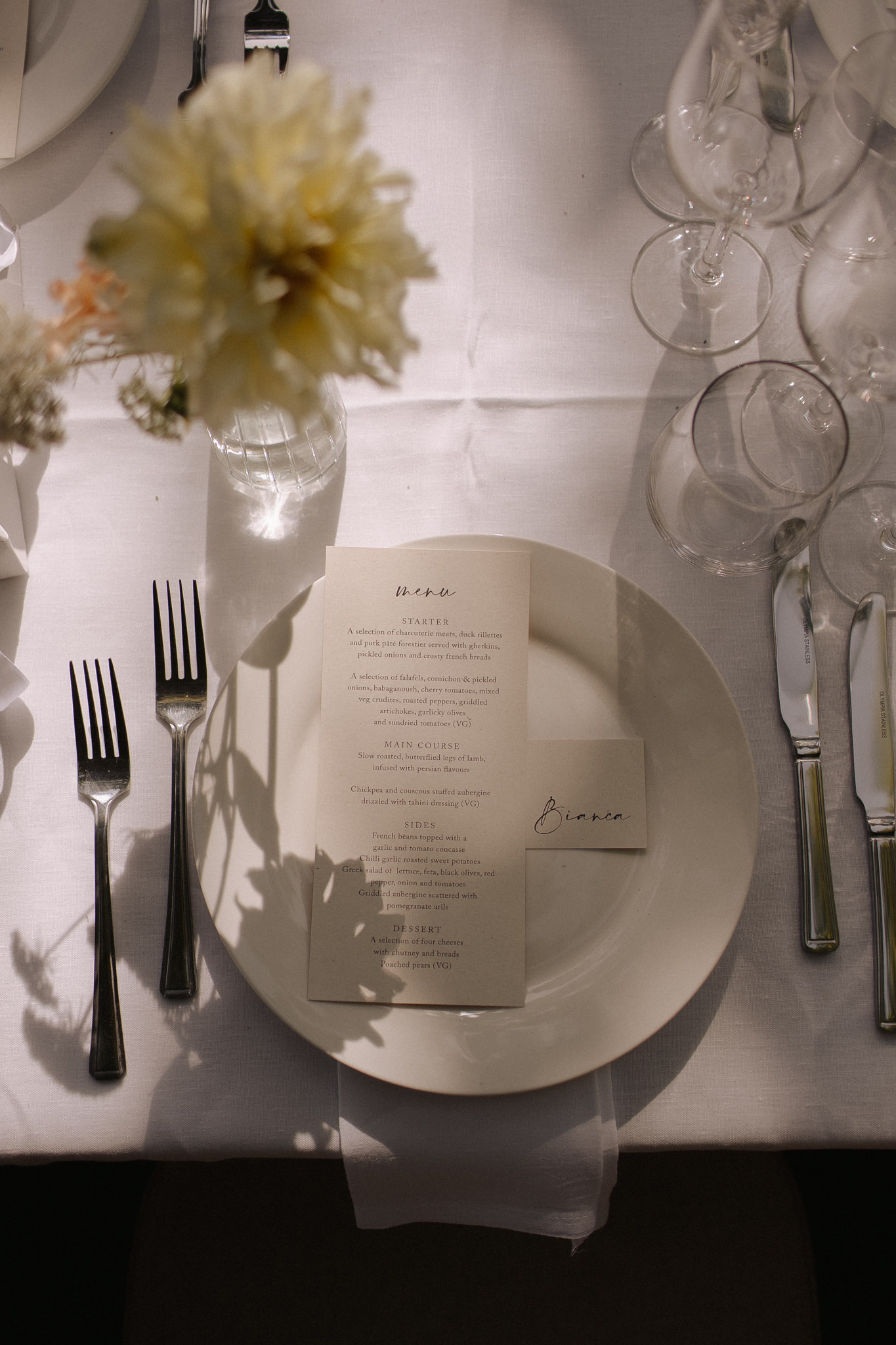 A close-up detail shot of a wedding reception place setting on a white linen tablecloth. The setting includes a white charger plate topped with a cream-toned printed menu card in a modern minimal typeface and a small handwritten place card reading 'Bianca'. Silver cutlery is arranged on either side — two forks to the left and a knife and butter knife to the right — alongside multiple clear wine and water glasses grouped to the upper right. A cream or pale yellow dahlia bloom in a clear glass bud vase is partially visible in the upper left, along with a soft peach dried floral stem, contributing to a neutral, pared-back decor palette. The overall styling is modern and minimal with a monochromatic cream and white color scheme.