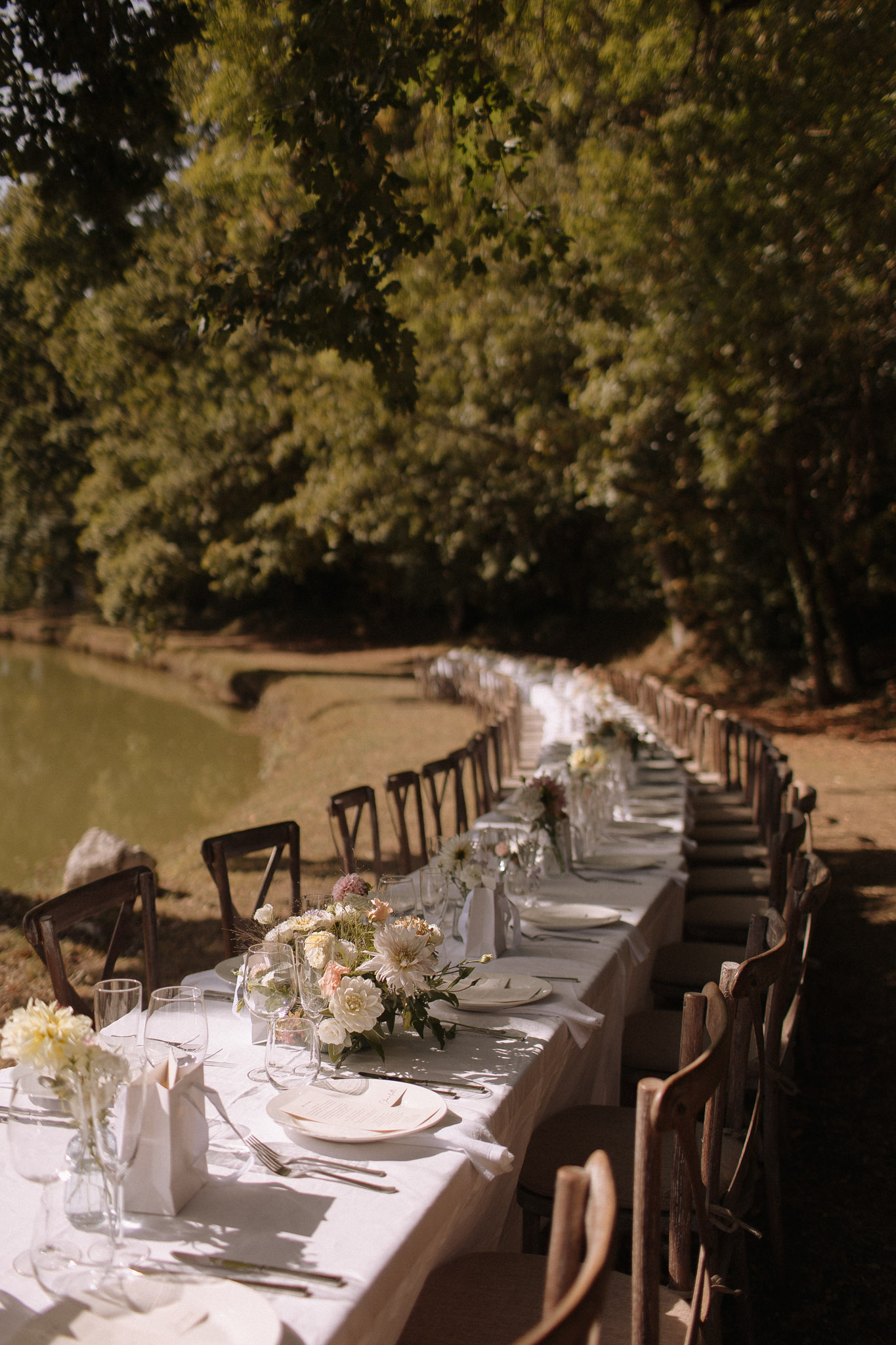 An outdoor wedding reception featuring a single long banquet table set alongside a small pond or lake, photographed from one end in a wide perspective shot. The table is dressed with a white linen tablecloth and set with white plates on blush chargers, folded white napkins, clear glassware including wine and water glasses, and silver cutlery. Floral arrangements run along the center of the table, featuring ivory ranunculus, blush and peach dahlias, and soft mauve blooms with green foliage, interspersed with small bud vases holding delicate white flowers. Dark wood cross-back chairs line both sides of the table, which extends far into the background. The overall styling is relaxed and organic with a neutral, earthy palette of white, blush, and cream tones, consistent with a natural or garden-inspired aesthetic.