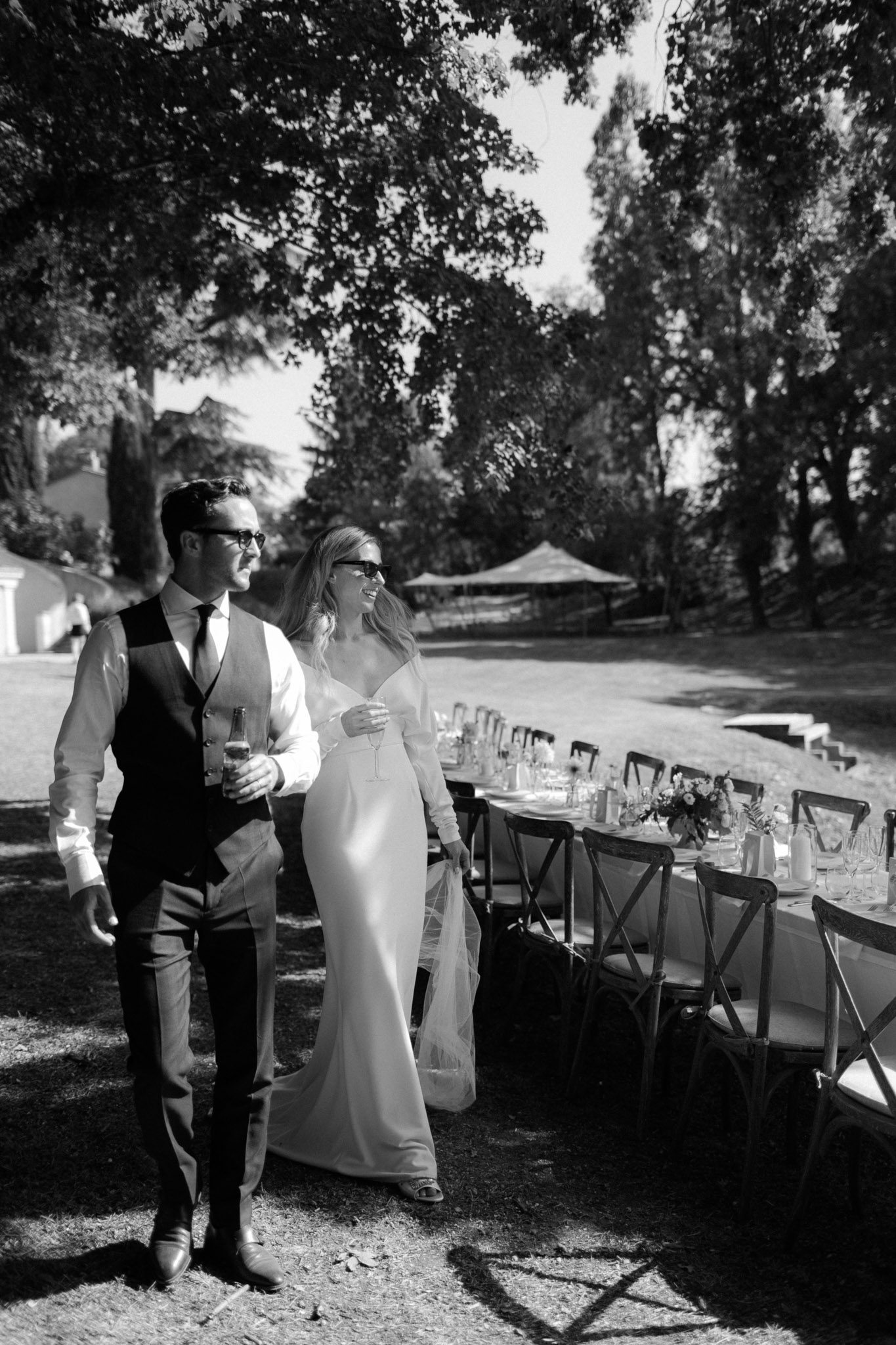 Black and white bride and groom in sunglasses walking along banquet table with champagne bottle
