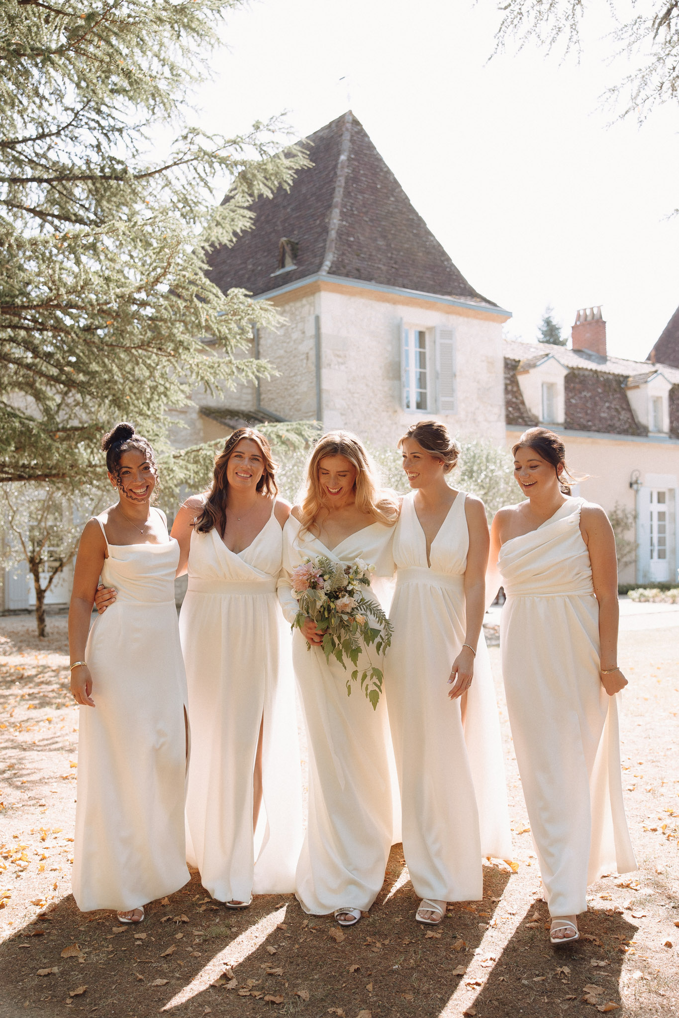 Bride and four bridesmaids in mismatched ivory dresses walking and laughing before stone chateau