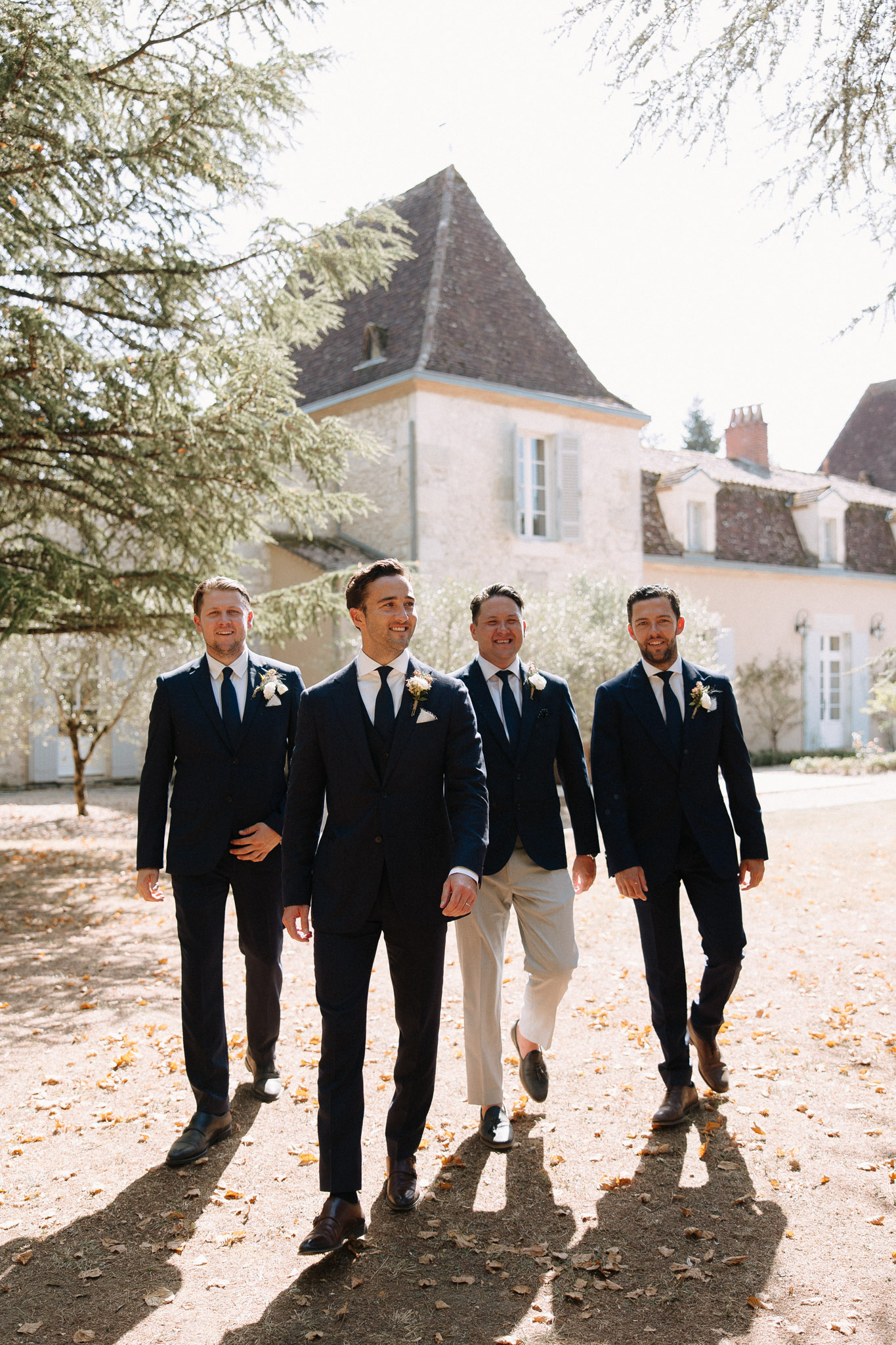 A groom and three groomsmen walk together across a gravel courtyard in front of a French château with a steep slate roof and stone facade. All four men wear navy suits with white dress shirts and navy ties, each with a small white boutonniere; one groomsman in the center-back stands out slightly with beige trousers rather than a full matching suit. The shot is taken in natural daylight with fallen autumn leaves on the ground, captured as a full-length walking portrait with the château prominently visible in the background.