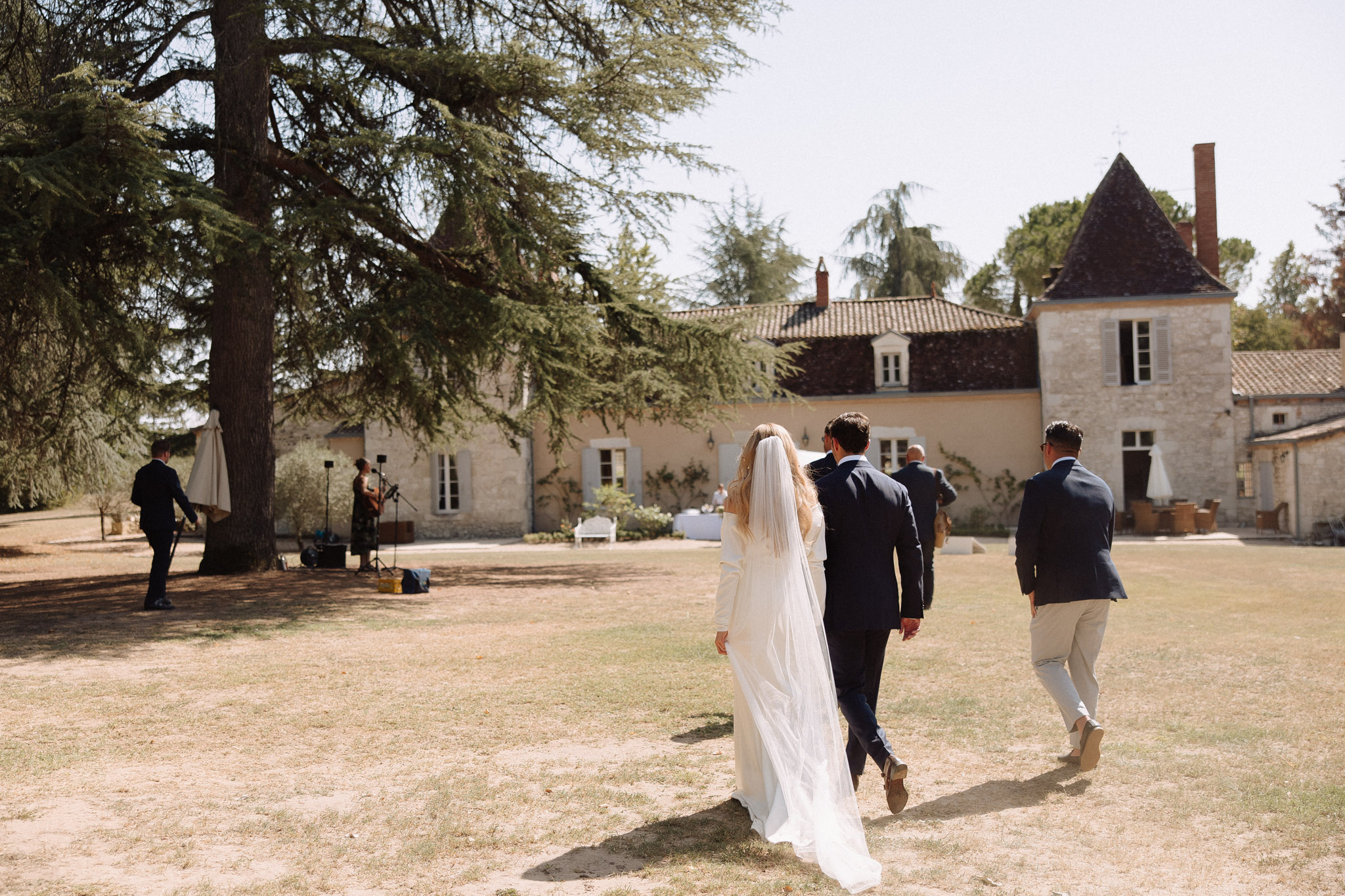 Bride and groom walking across chateau grounds from behind, musician performing near stone tower facade