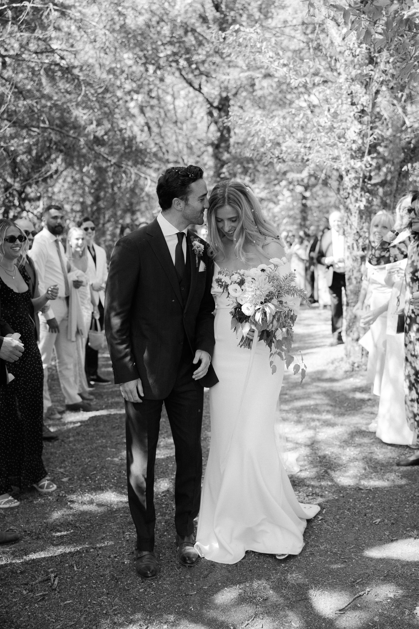 This black-and-white image captures the couple walking back up the aisle immediately after an outdoor ceremony, surrounded by approximately 20–30 guests lined on either side of a tree-lined path. The groom wears a dark fitted suit with a dark tie, pocket square, and a small boutonnière, and leans toward the bride with a smile. The bride wears a slim, off-the-shoulder gown with a slight train and carries a full, loosely arranged bouquet featuring large blooms — identifiable as ranunculus or anemones — with trailing foliage. The image has high contrast with bright highlights from dappled sunlight filtering through the tree canopy above. The composition is a mid-length portrait shot taken at eye level, with the couple sharp in the foreground and guests softly visible in the background.