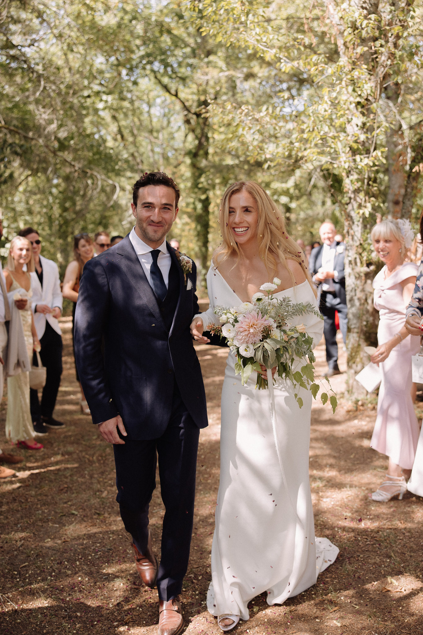 The bride and groom walk back up the aisle together immediately after their ceremony, outdoors along a tree-lined dirt path with guests lining both sides. The groom wears a navy suit with a white shirt, dark navy tie, and tan leather Oxford shoes, with a small floral buttonhole. The bride wears an off-shoulder white satin slip-style gown with a short train and carries a loose, garden-style bouquet featuring blush dahlias, white ranunculus, and trailing eucalyptus and greenery. Guests in the background are smiling and appear to be throwing confetti, with visible attire including blush, white, and dark formal wear. The shot is a medium full-length portrait taken head-on, with natural dappled light filtering through the tree canopy overhead.